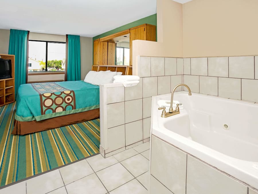 A hotel room with a bed, a large window, a television, a bathtub, and a wooden wardrobe.