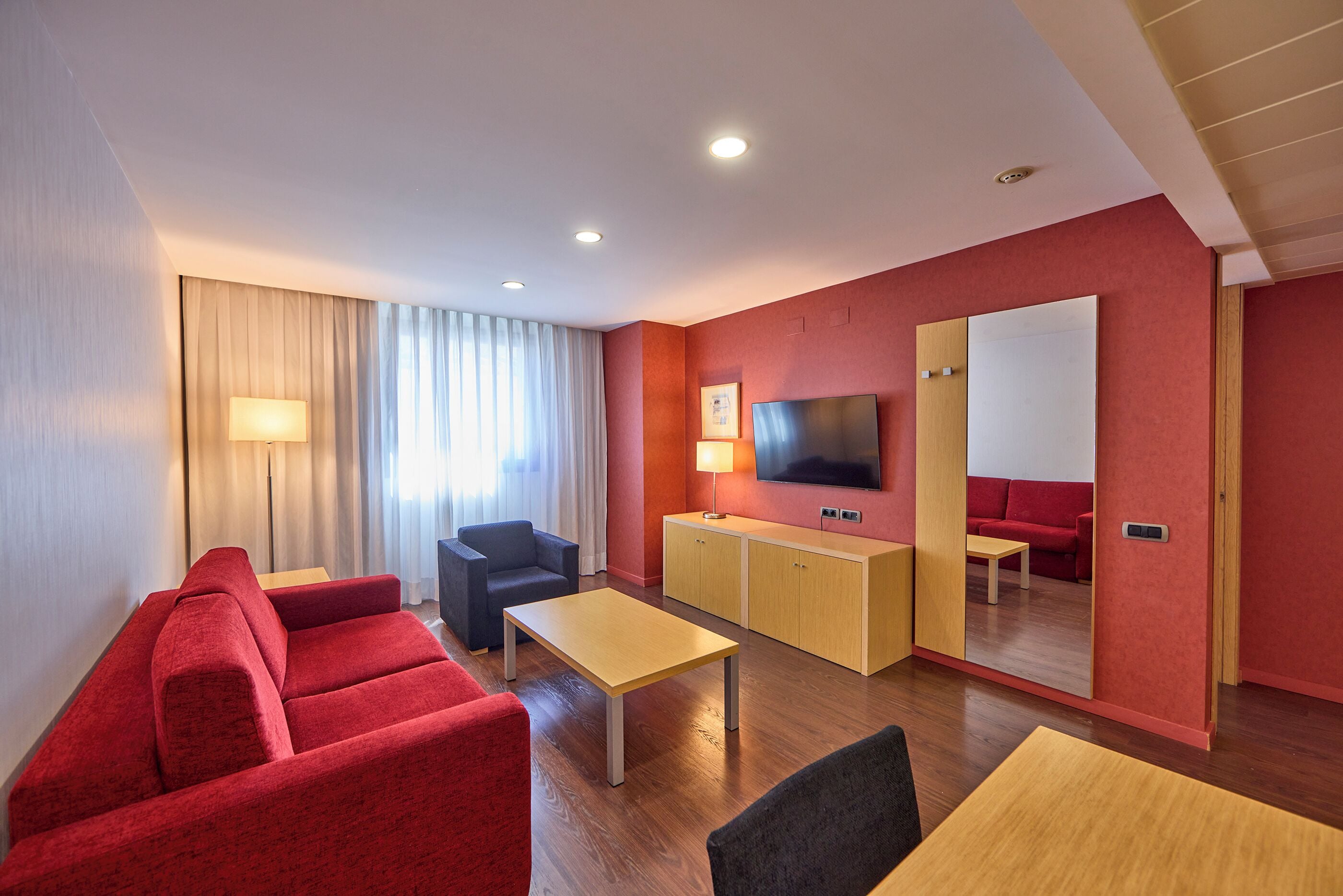Une chambre d’hôtel moderne avec un canapé rouge, une petite table, un téléviseur et un miroir.