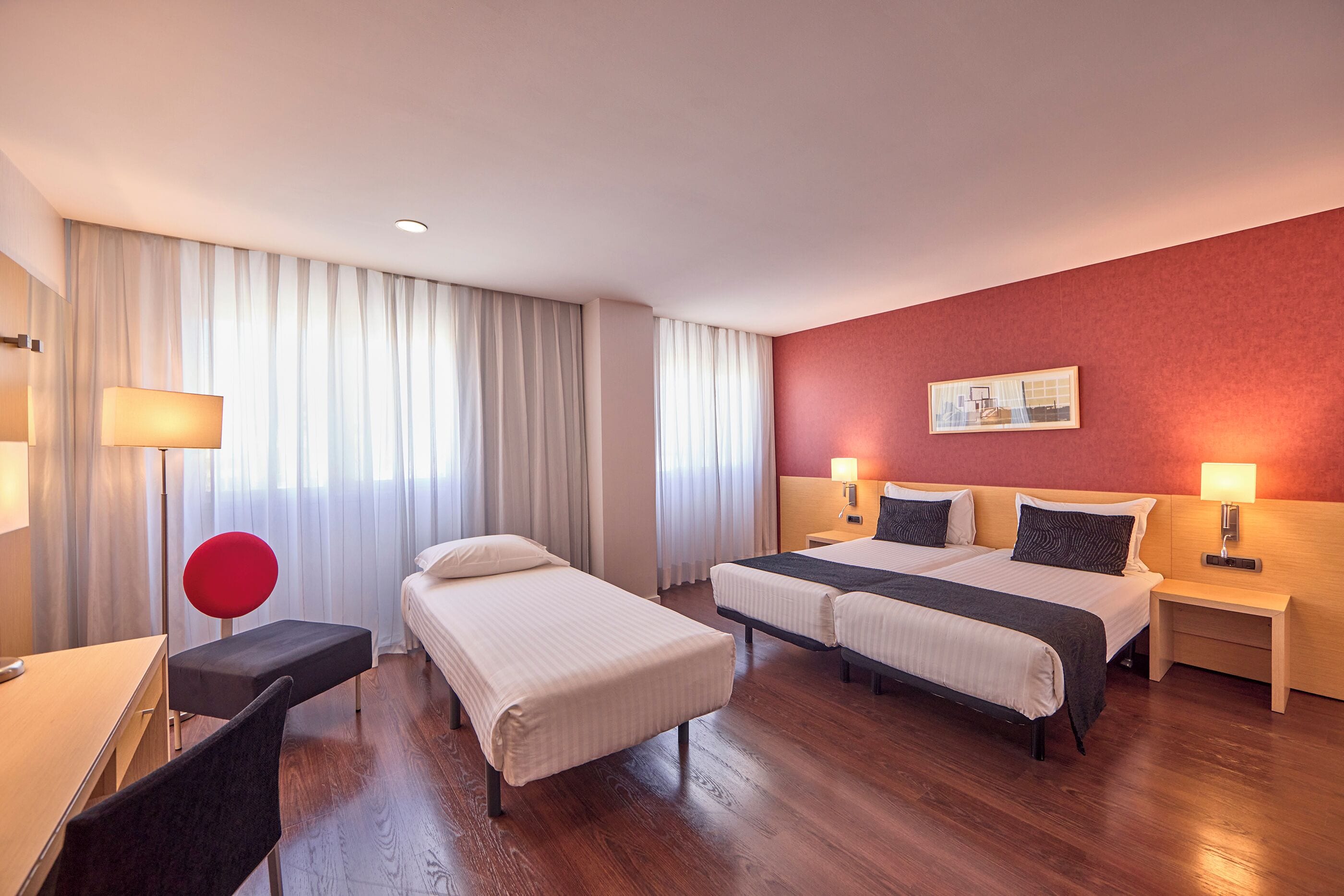 A hotel room with two single beds, a desk, a chair, a red chair, a bedside table with a lamp, and a large window with curtains.
