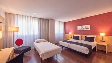 A hotel room with two single beds, a desk, a chair, a red chair, a bedside table with a lamp, and a large window with curtains.