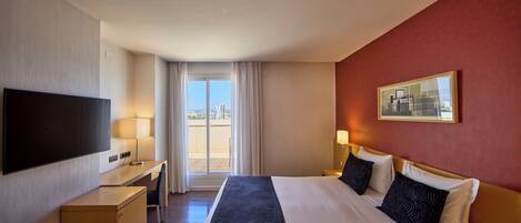 A hotel room with a bed, desk, TV, and a view of the outdoors.