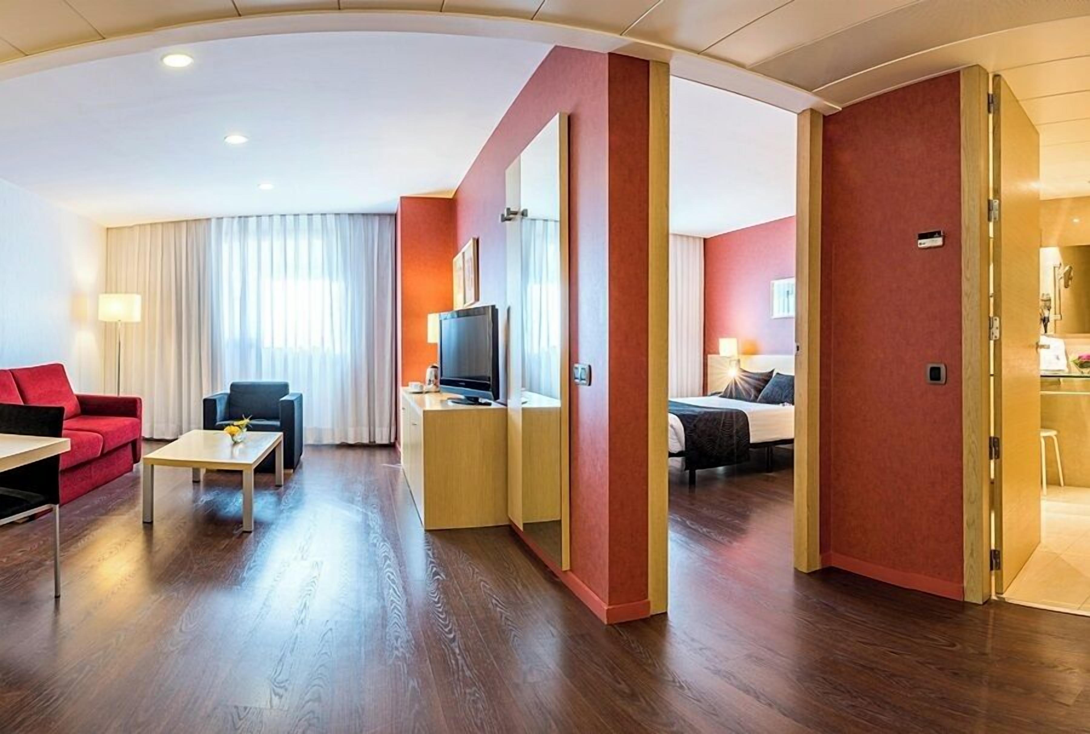 Habitació d'hotel amb sòl de fusta, un sofà vermell, una taula petita, televisió, llit i un bany visible a través d'una porta oberta.