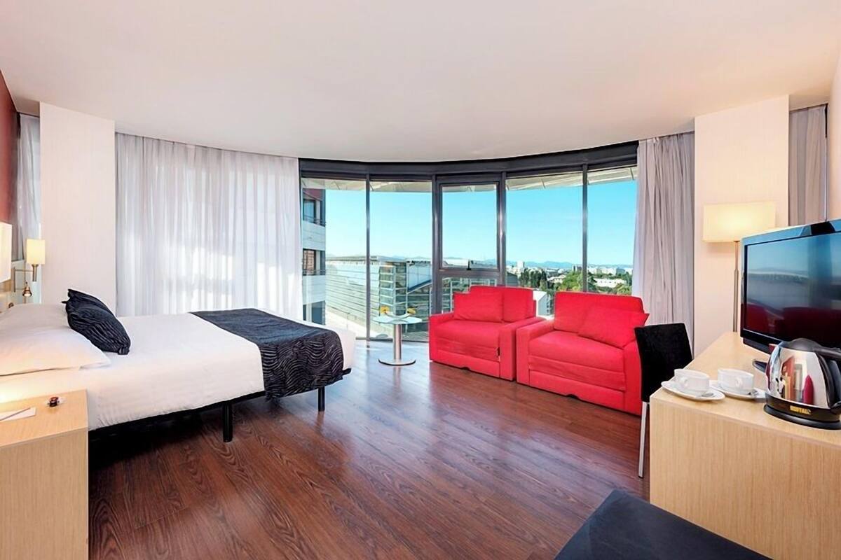 A hotel room with a bed, a red sofa, a TV, and a view of the city.