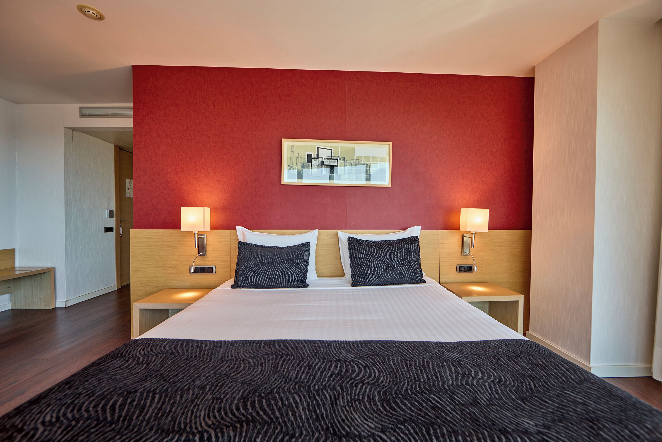 A hotel room with a large bed, two bedside tables with lamps, a red accent wall, and a framed picture above the bed.