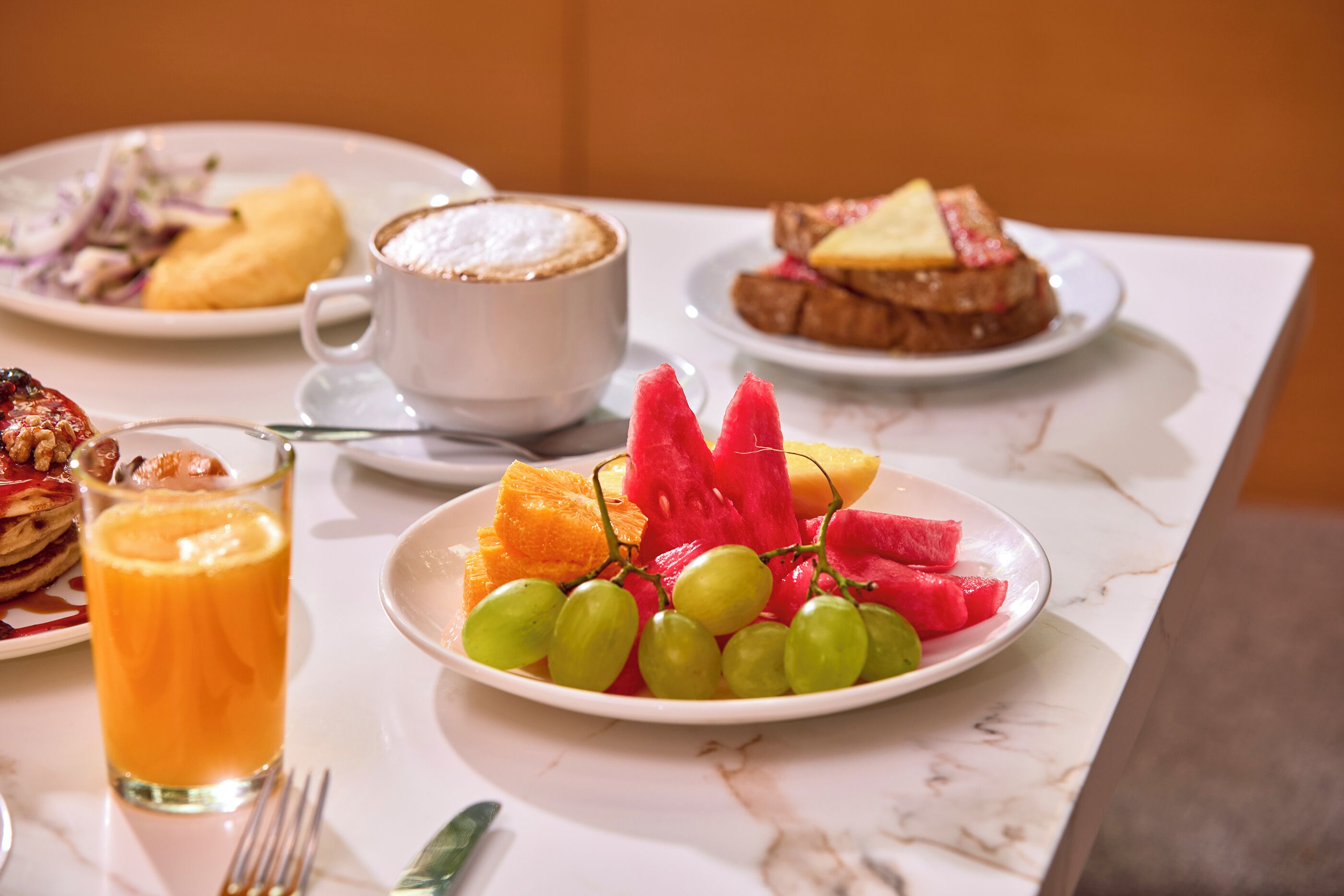 Une table en marbre dressée pour le petit-déjeuner, avec des assiettes de fruits, un verre de jus d’orange et une tasse de café.