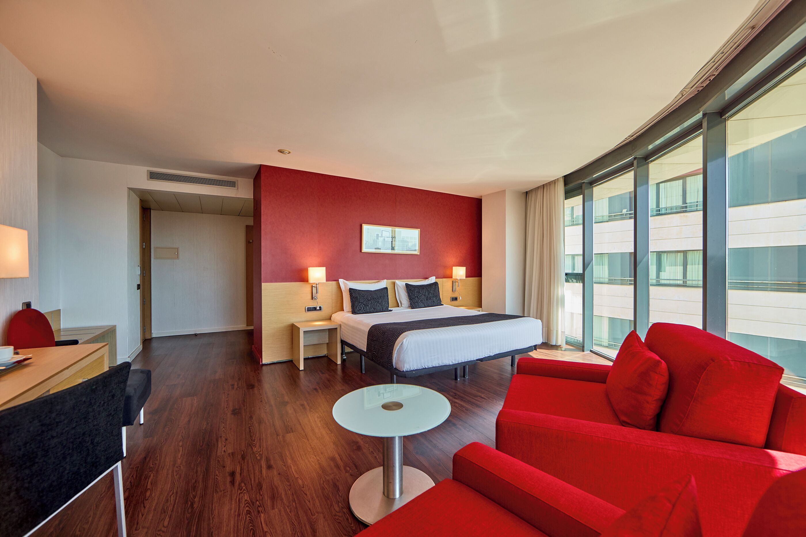 A hotel room with a bed, a red sofa, a small table, and a desk with a lamp.