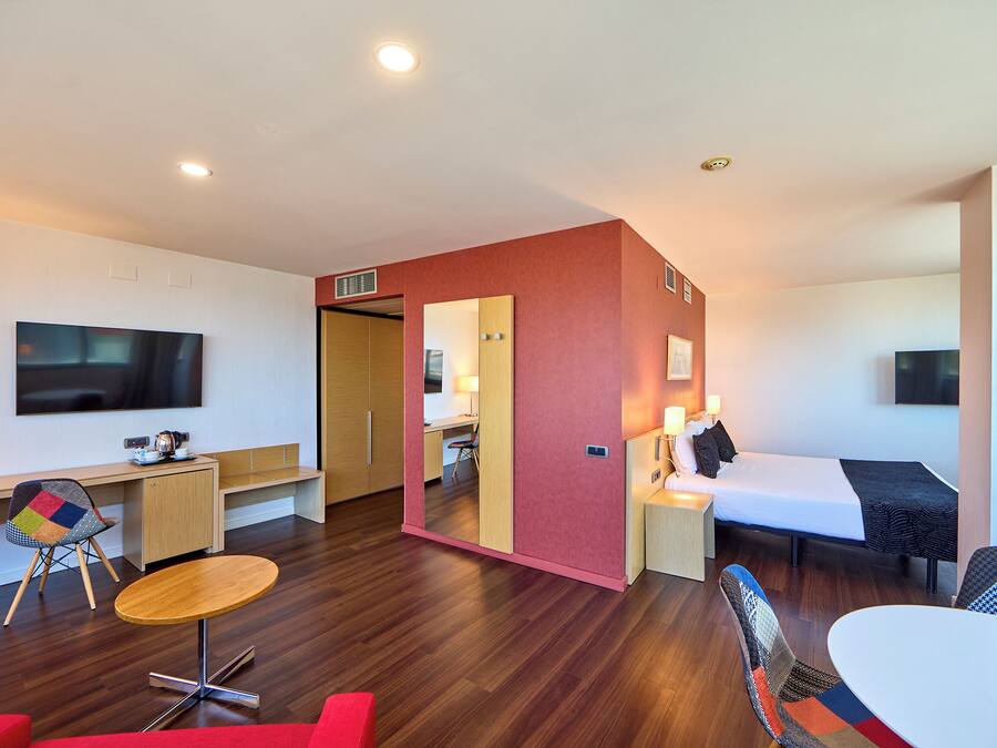 A modern hotel room with a wooden desk, a TV, a bed, and a small dining area.