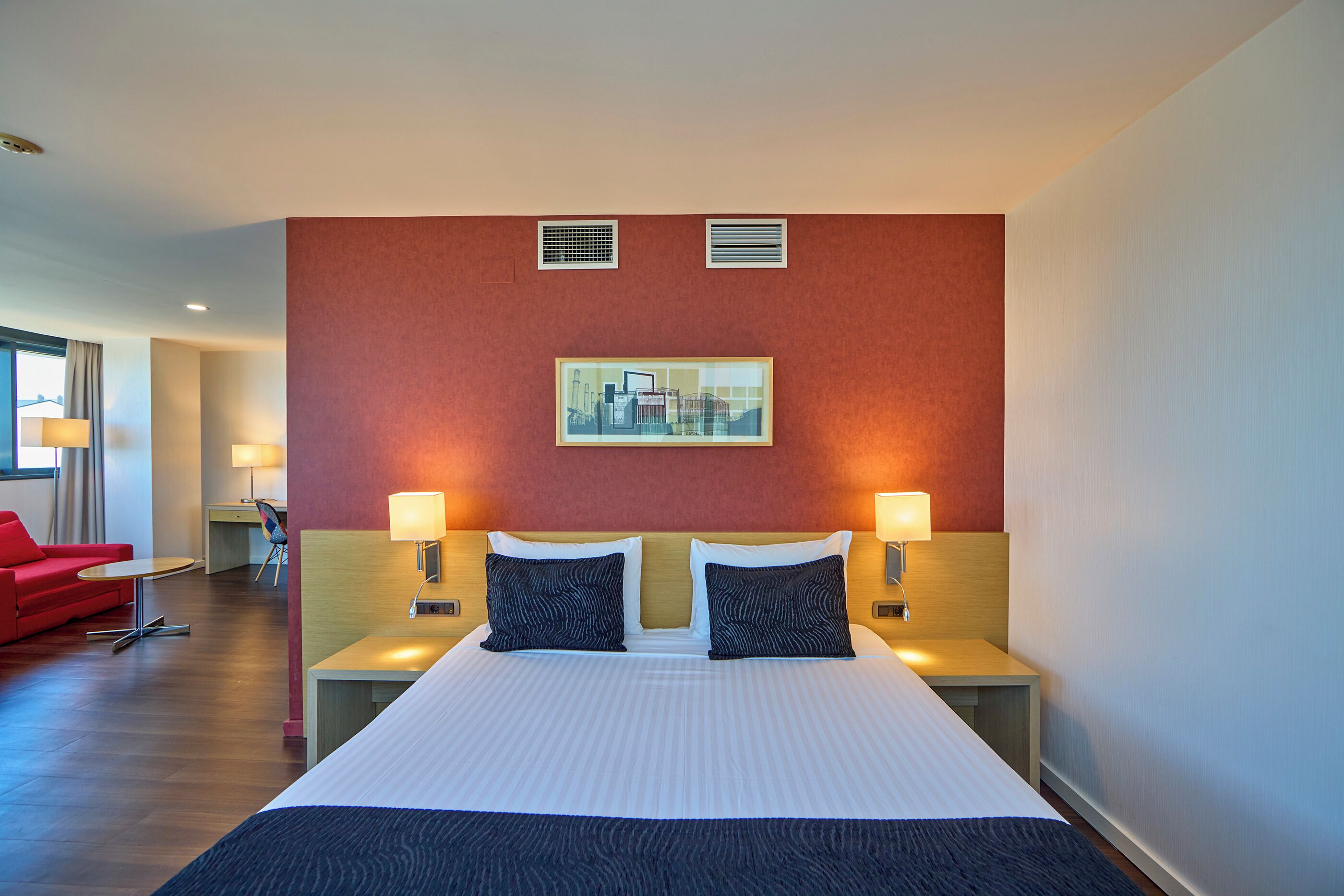 A hotel room with a large bed, two bedside tables with lamps, a red sofa, a desk, and a cityscape painting on the wall.