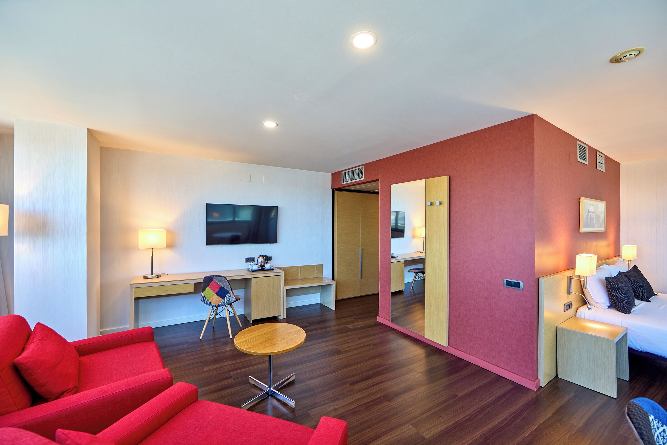 A modern hotel room with a red sofa, a small round table, a desk with a chair, a bed with a nightstand, and a large mirror.