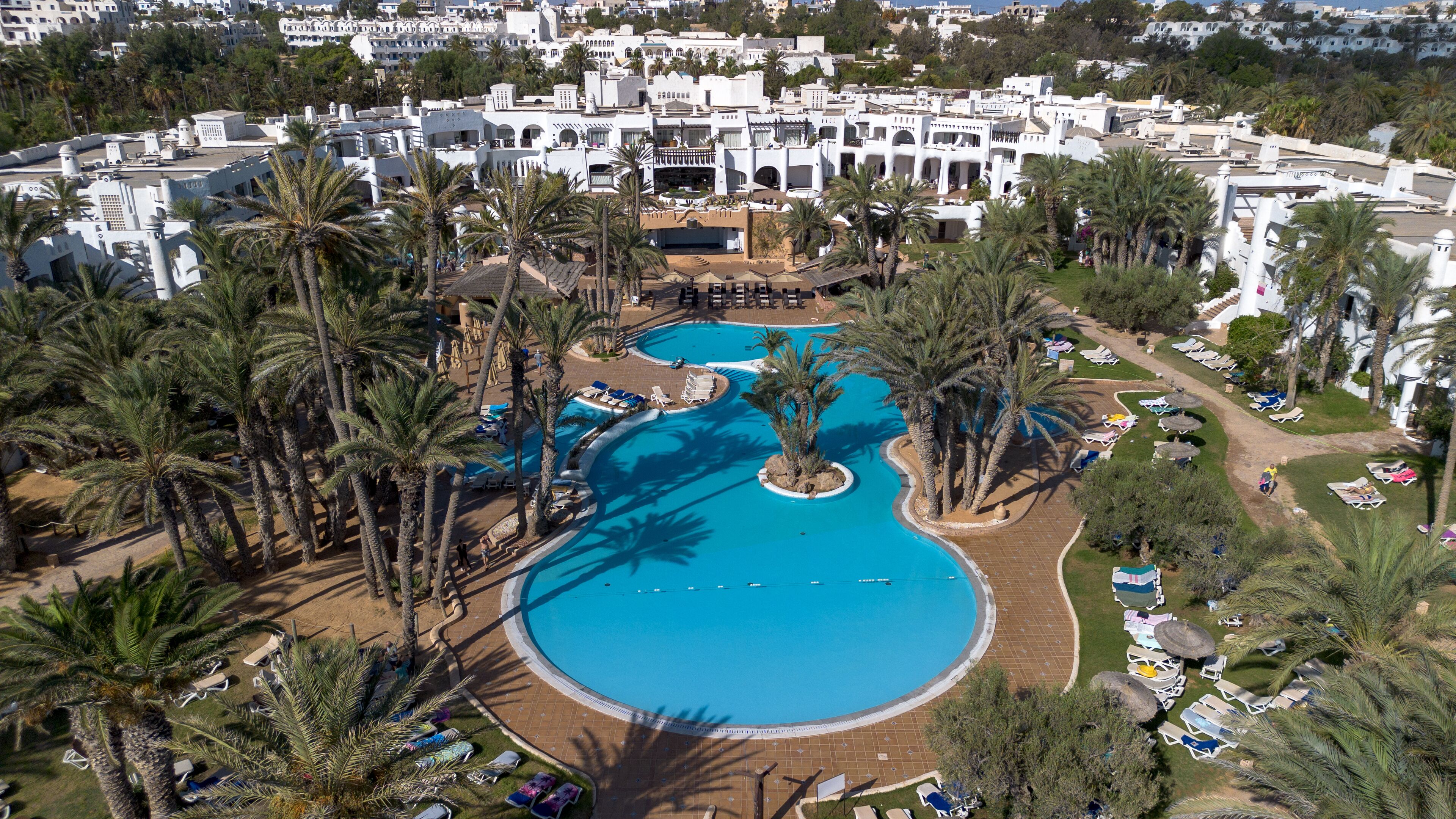 A resort with a large central pool surrounded by palm trees and sun loungers.