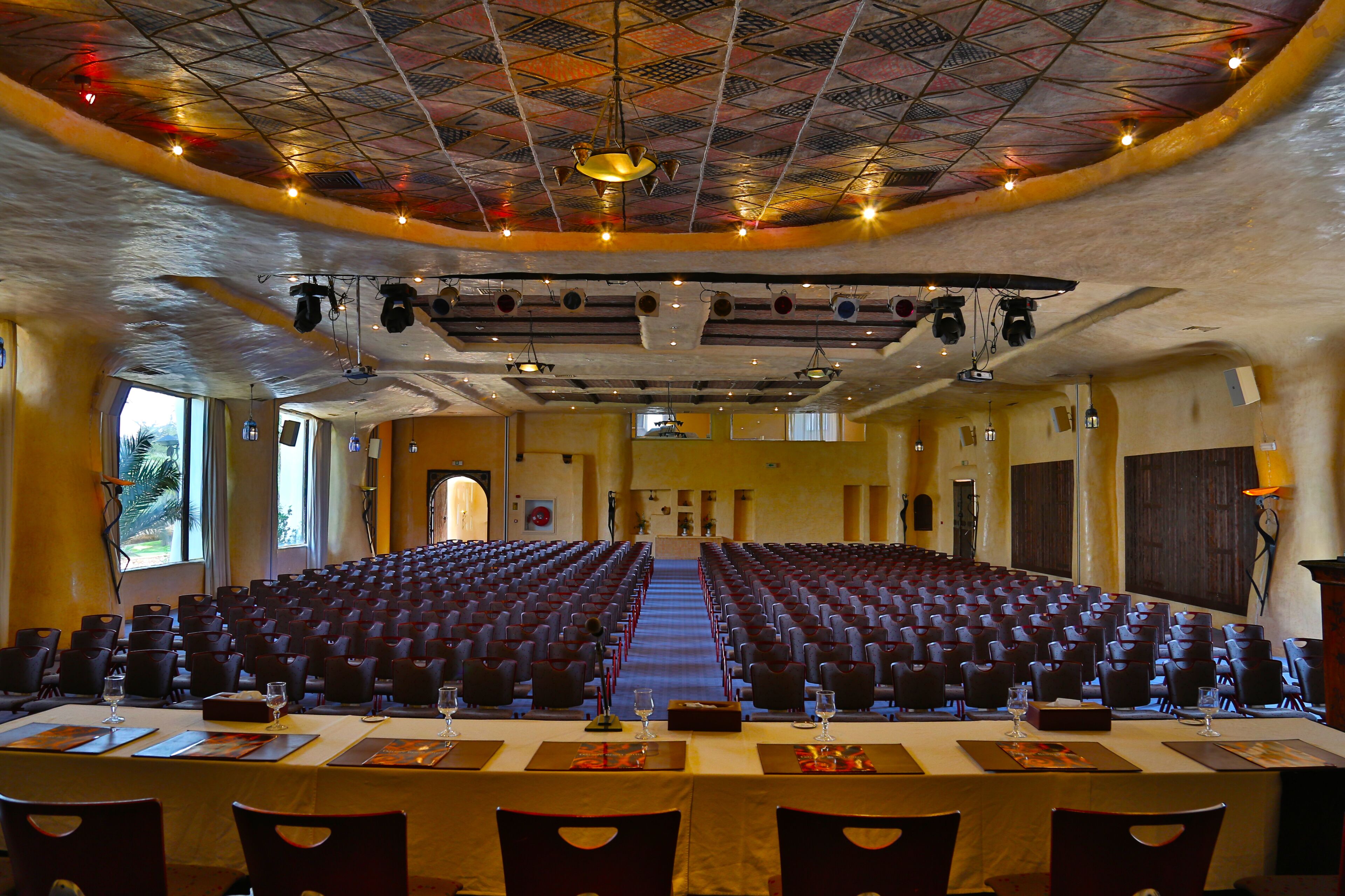 A conference room with rows of chairs, a long table with placemats and glasses, and a patterned ceiling.