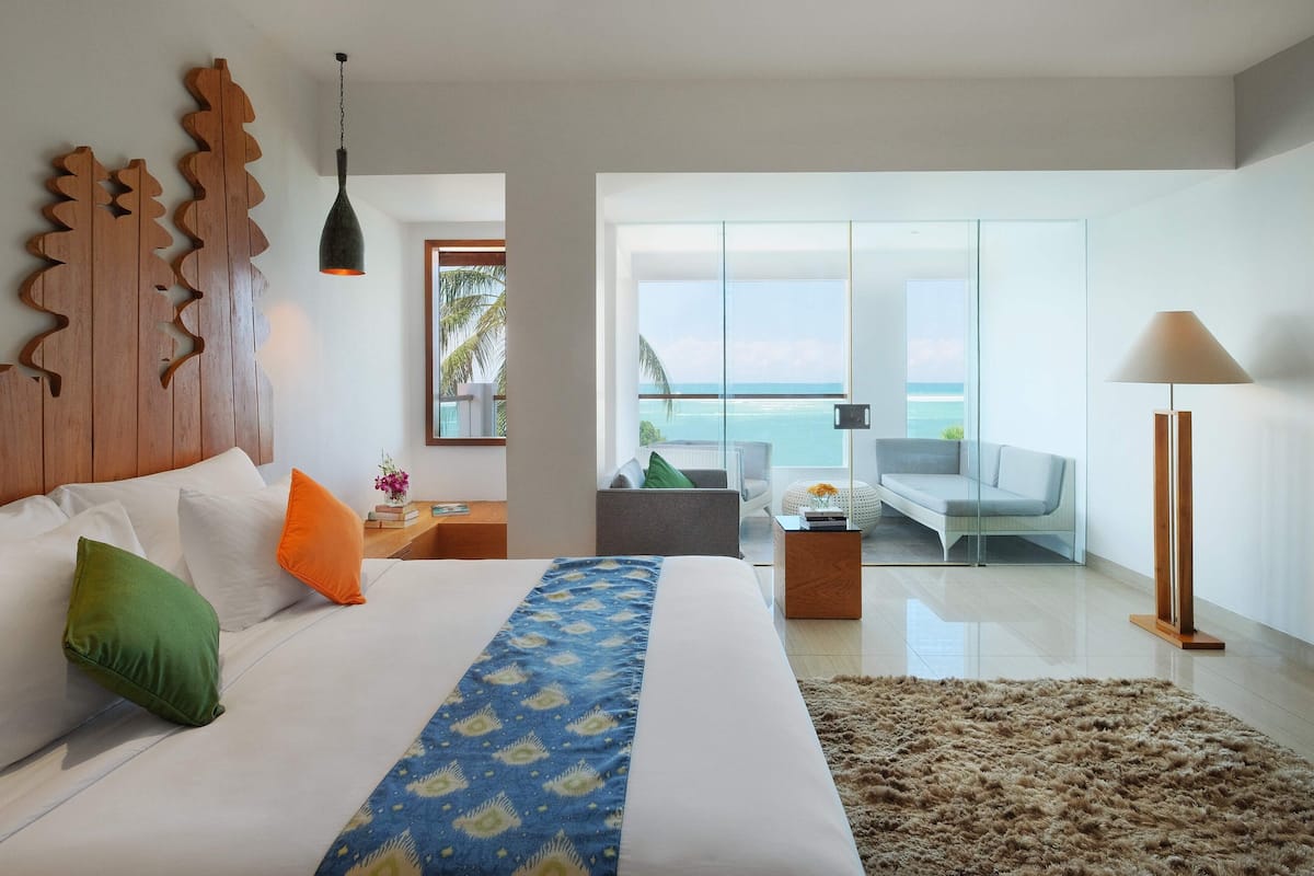 A modern hotel room with a large bed, a view of the ocean, a balcony with seating, and a wooden headboard.