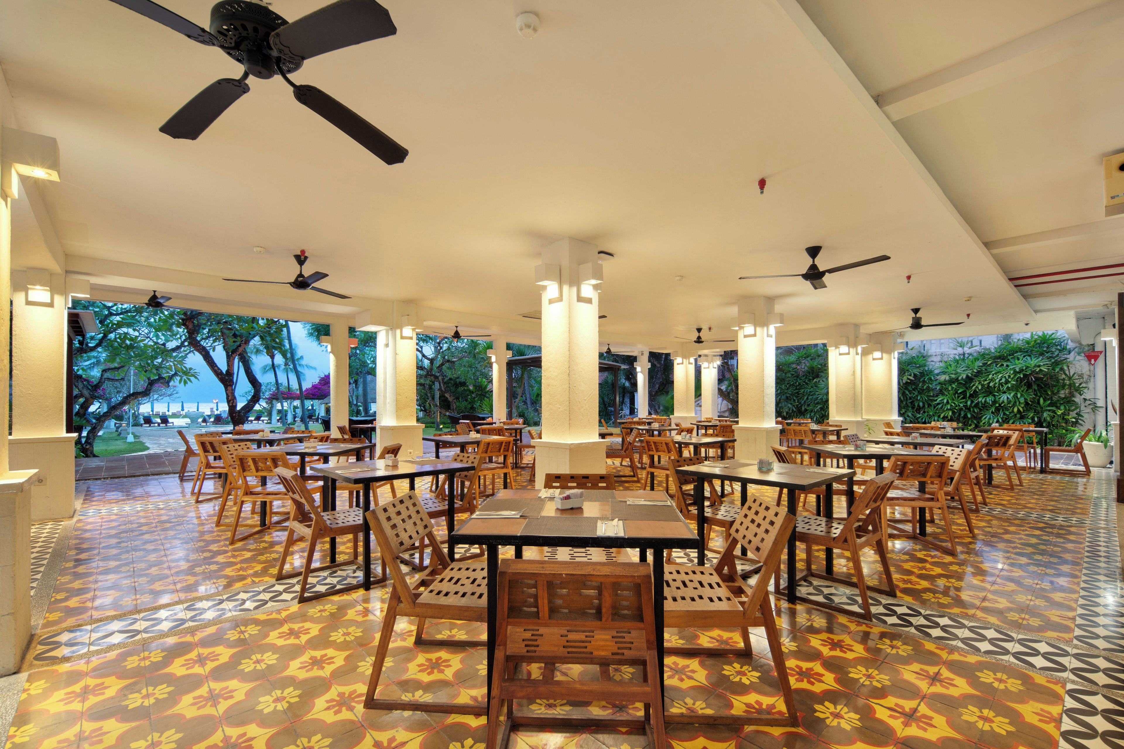 A spacious indoor dining area with multiple tables and chairs, ceiling fans, and large windows offering a view of the outdoors.