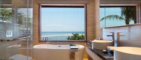 Een moderne badkamer met een groot bad, twee wastafels en een groot raam met uitzicht op het strand.