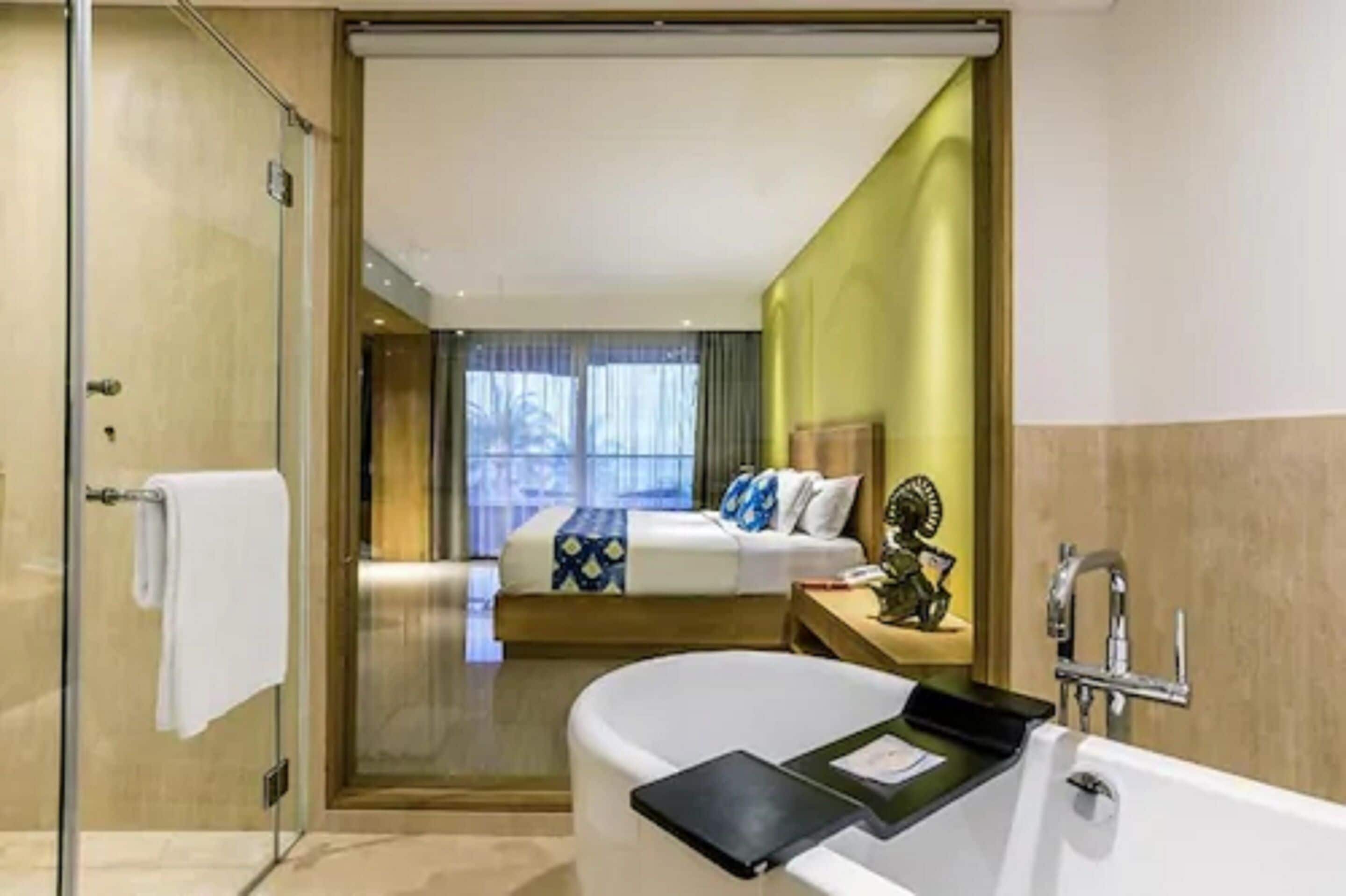 A hotel room with a glass shower enclosure, a large bathtub, a bed with a blue and white bedspread, and a view of the outdoors through the window.