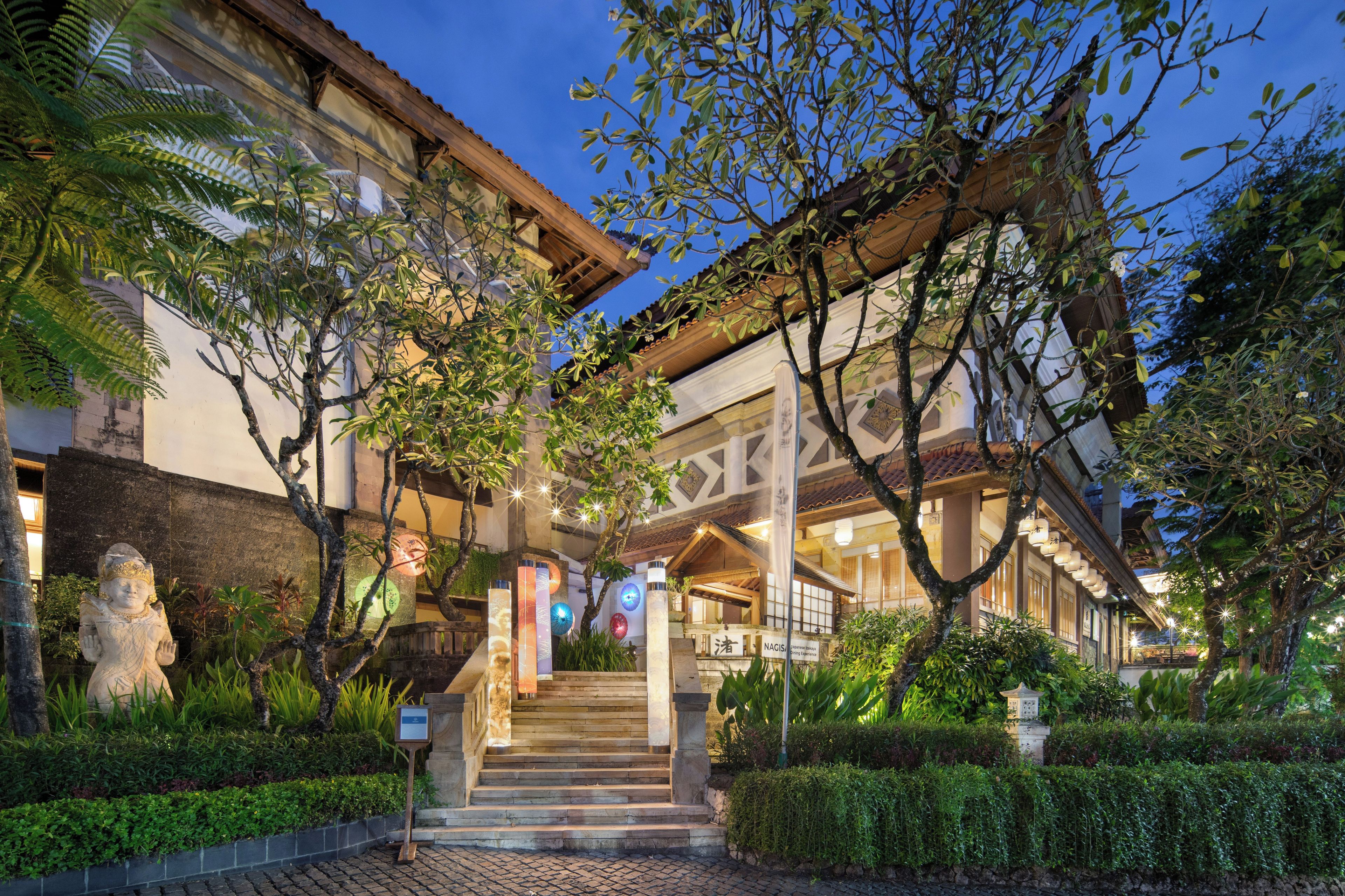 A traditional building with a staircase leading to a covered entrance, surrounded by lush greenery and stone statues.