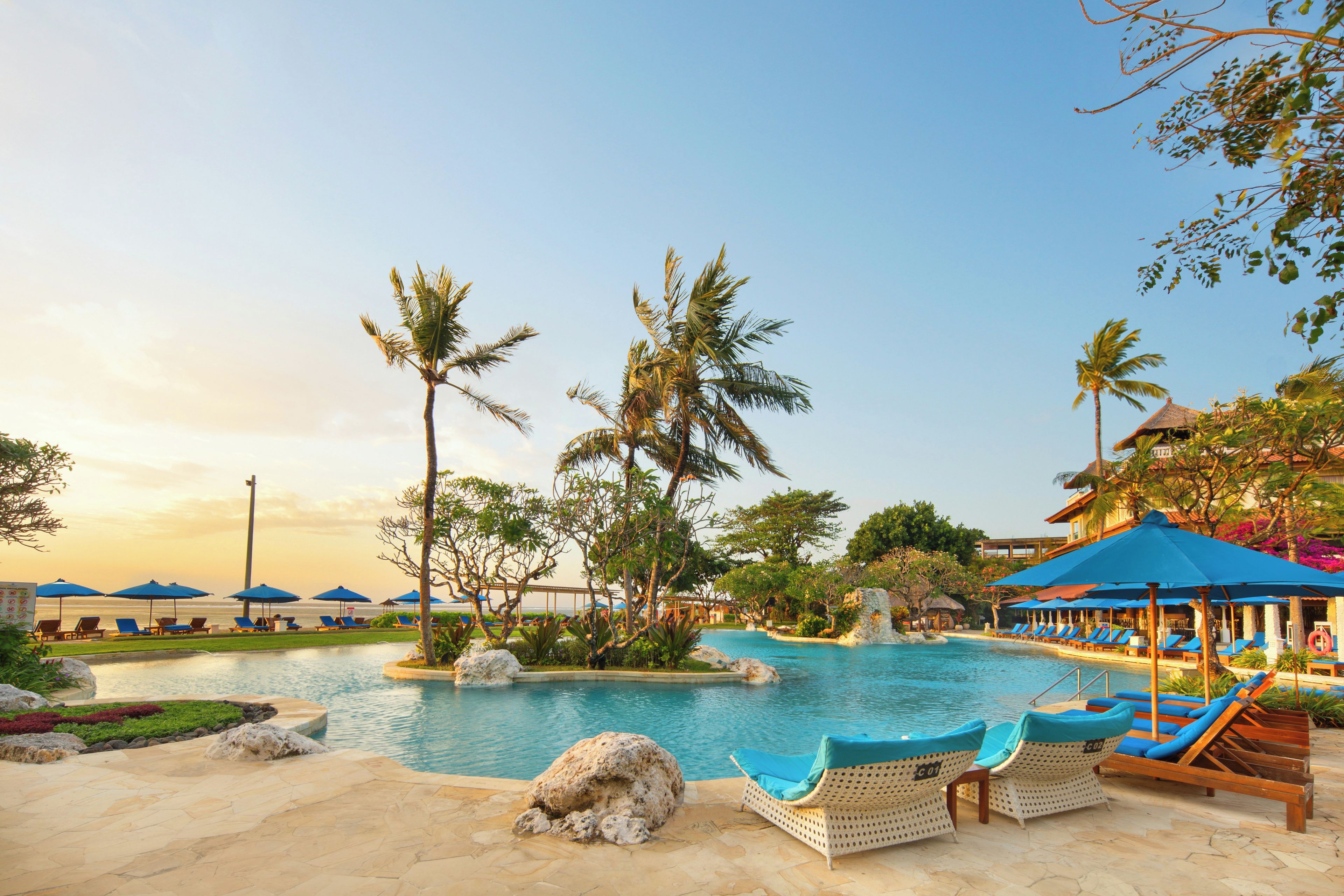 A pool area with lounge chairs and umbrellas, surrounded by palm trees and a clear sky.