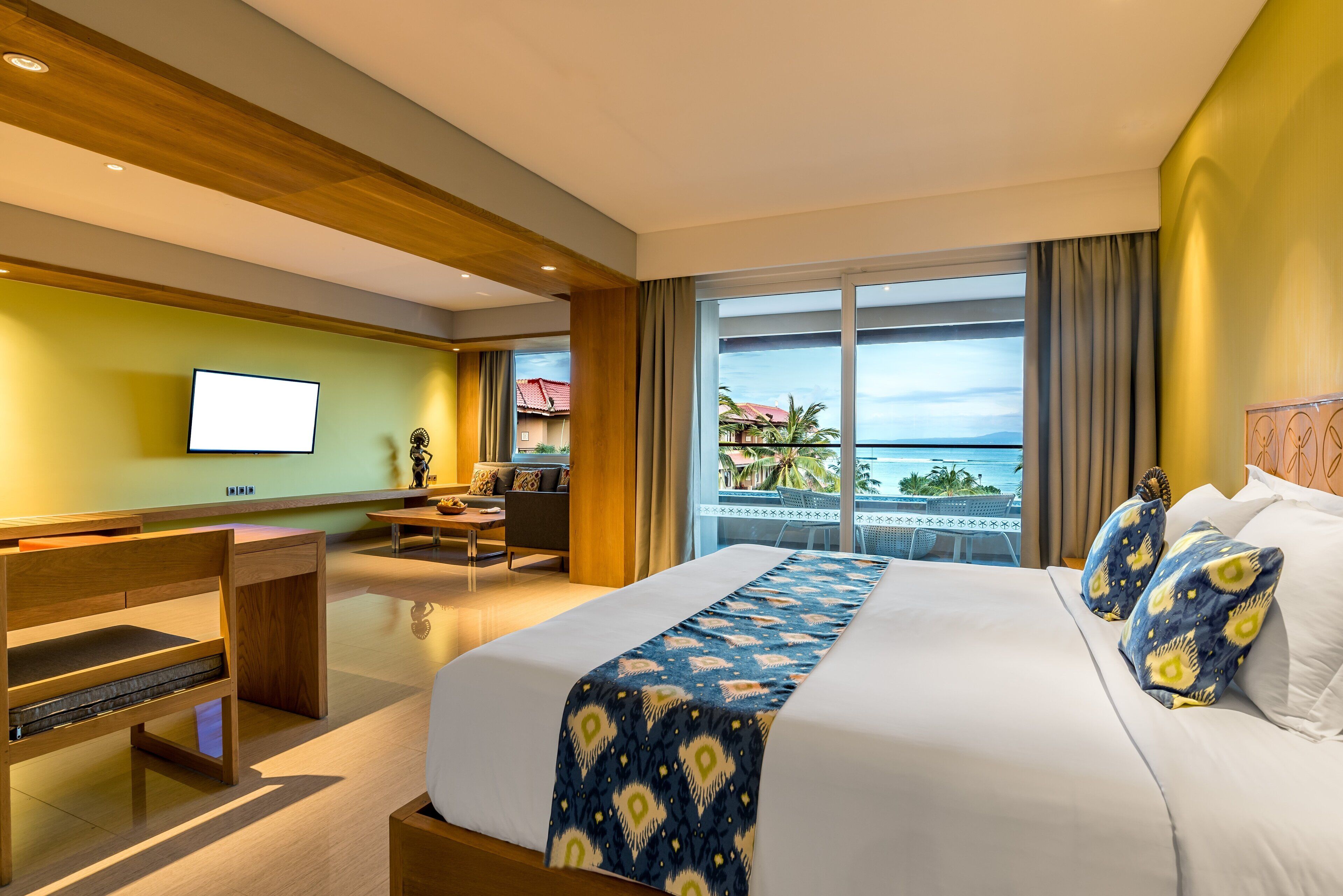A hotel room with a large bed, a desk, a TV, and a balcony with a view of the ocean.