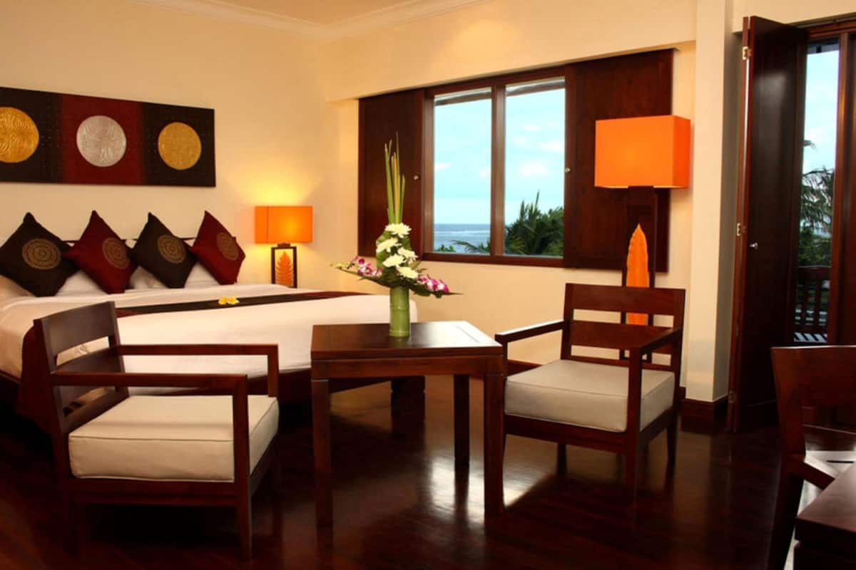 A hotel room with a bed, two chairs, a small table with a vase of flowers, a lamp, and a window with a view of the ocean.