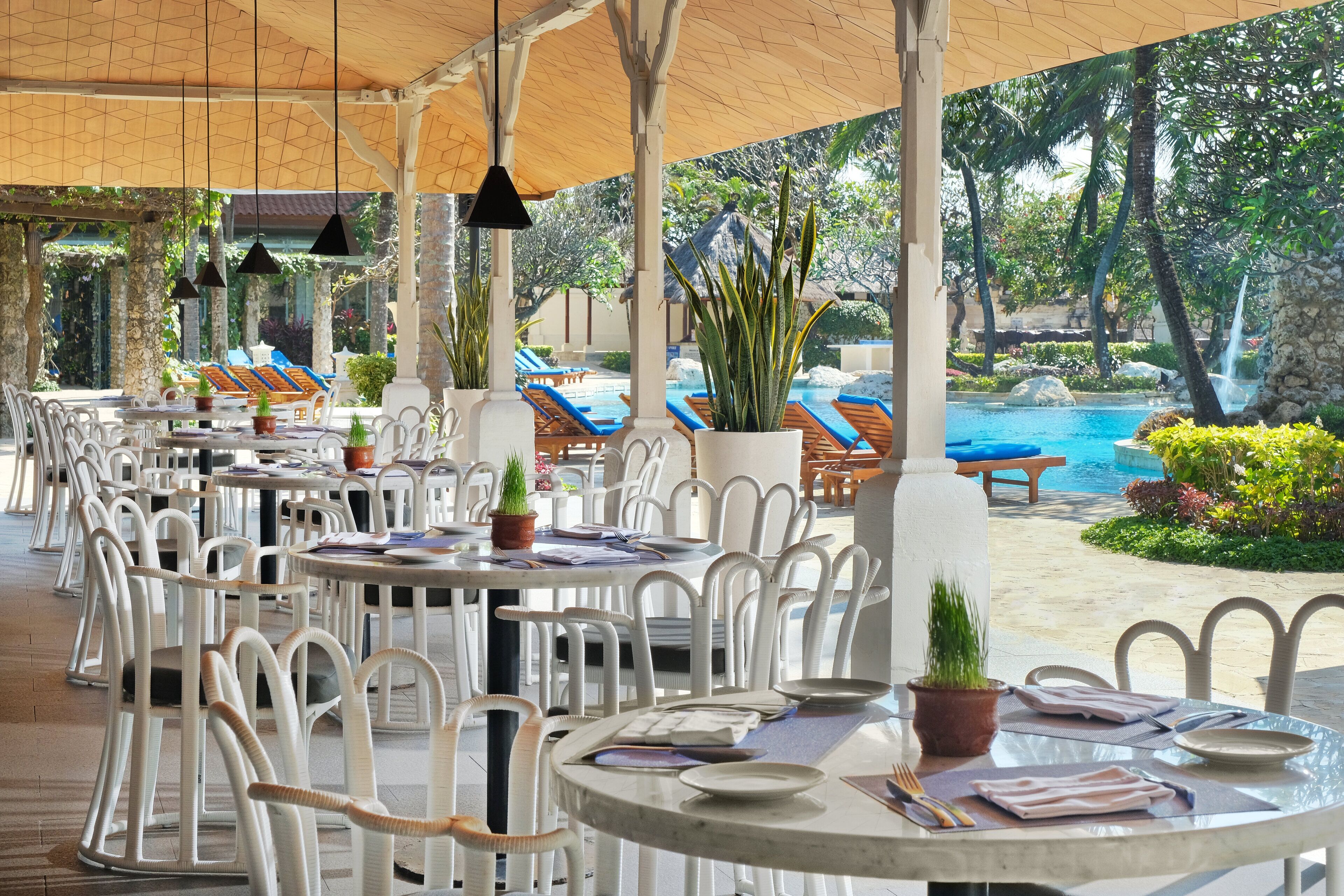 Un área de comedor al aire libre con techo, sillas y mesas blancas, una piscina y plantas tropicales.