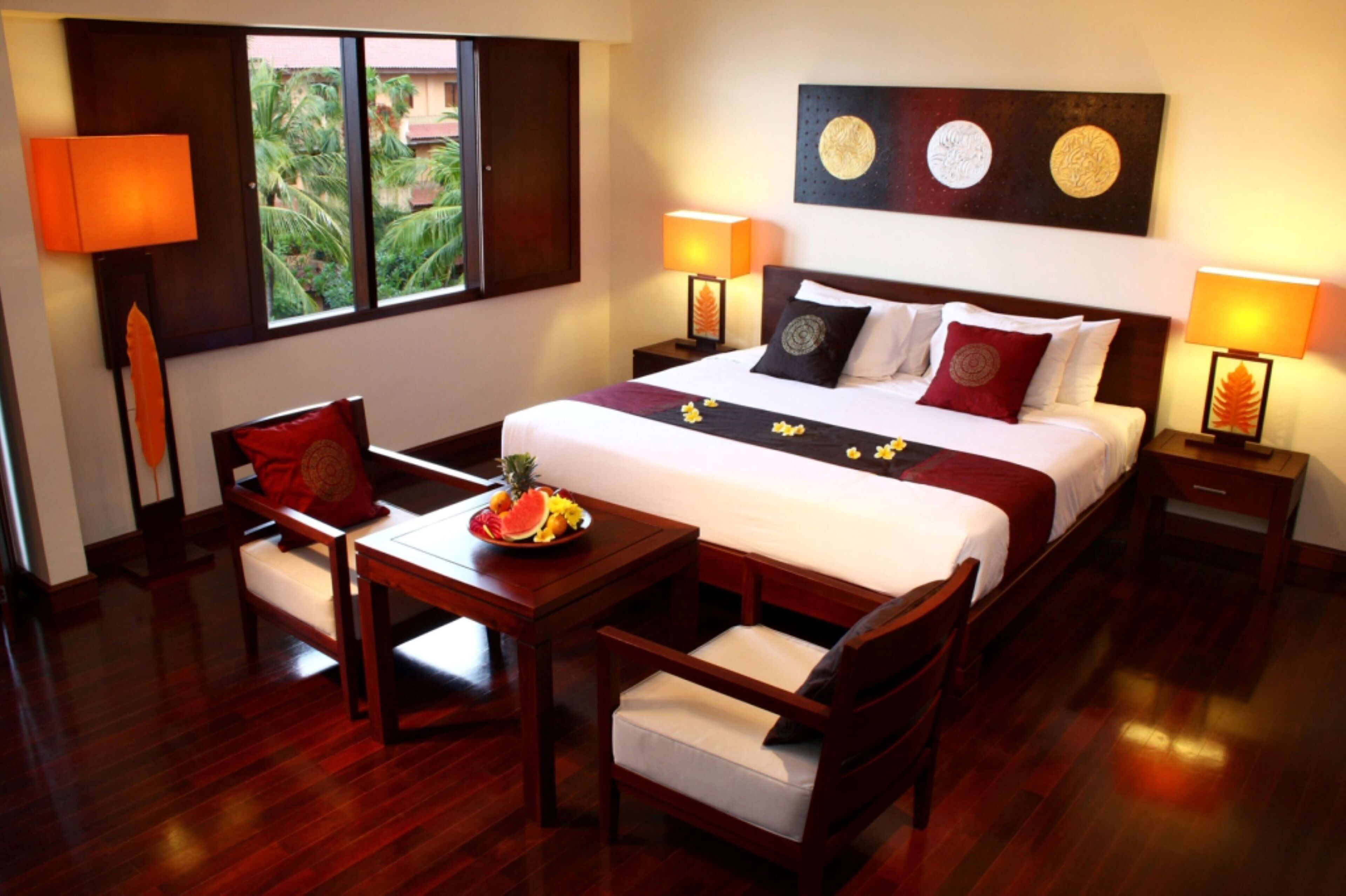 A hotel room with a bed, two bedside tables, a desk, two chairs, a window with a view of greenery, and wall art.