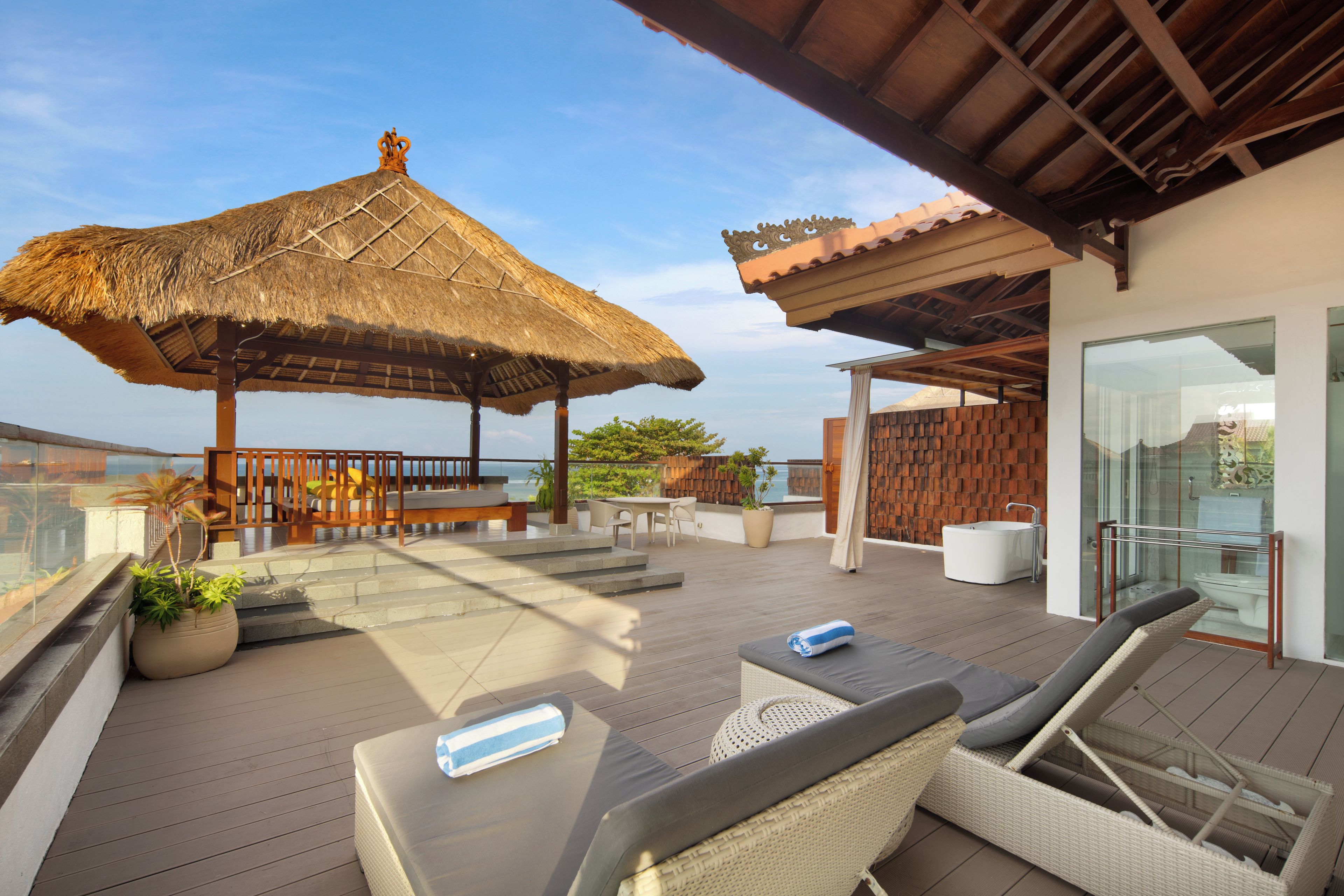 Una terraza en la azotea con sombrilla de paja, mobiliario exterior y un baño con paredes de cristal.