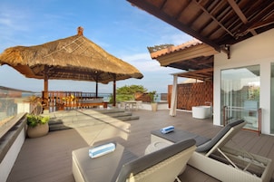 Una terraza en la azotea con sombrilla de paja, mobiliario exterior y un baño con paredes de cristal.