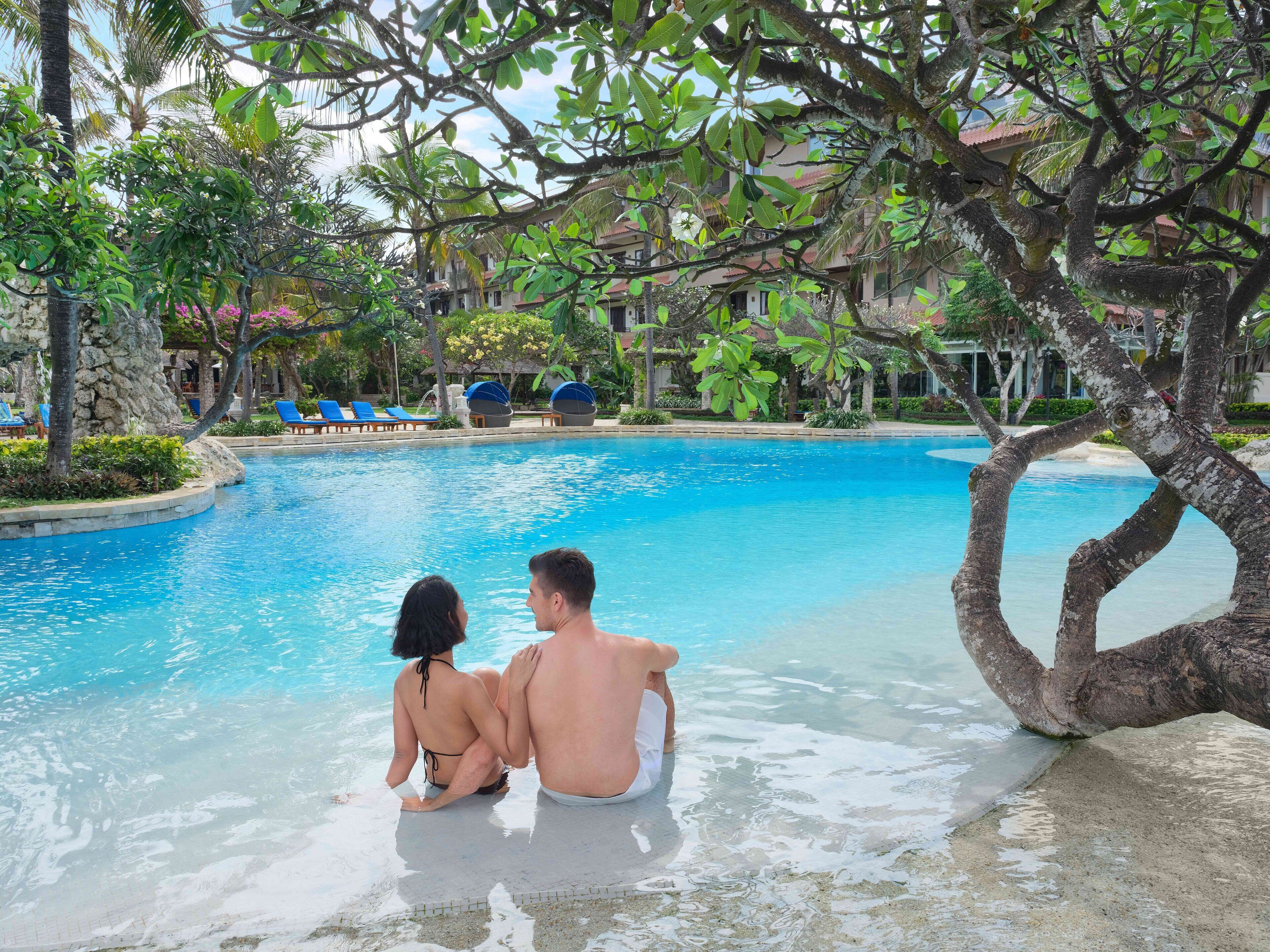 A couple sitting by a clear pool surrounded by lush greenery and a resort building in the background.