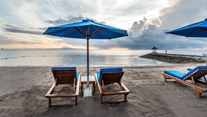 Una playa con sillones de madera y sombrillas azules, un rompeolas rocoso y un mar tranquilo bajo un cielo nublado.