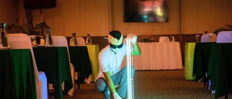 A person setting up a light fixture in a room with tables covered in green and white tablecloths, a flat-screen TV on the wall, and a carpeted floor.