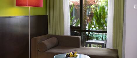 A room with a sofa, a glass table with fruit, a red lamp, and a view of greenery outside.