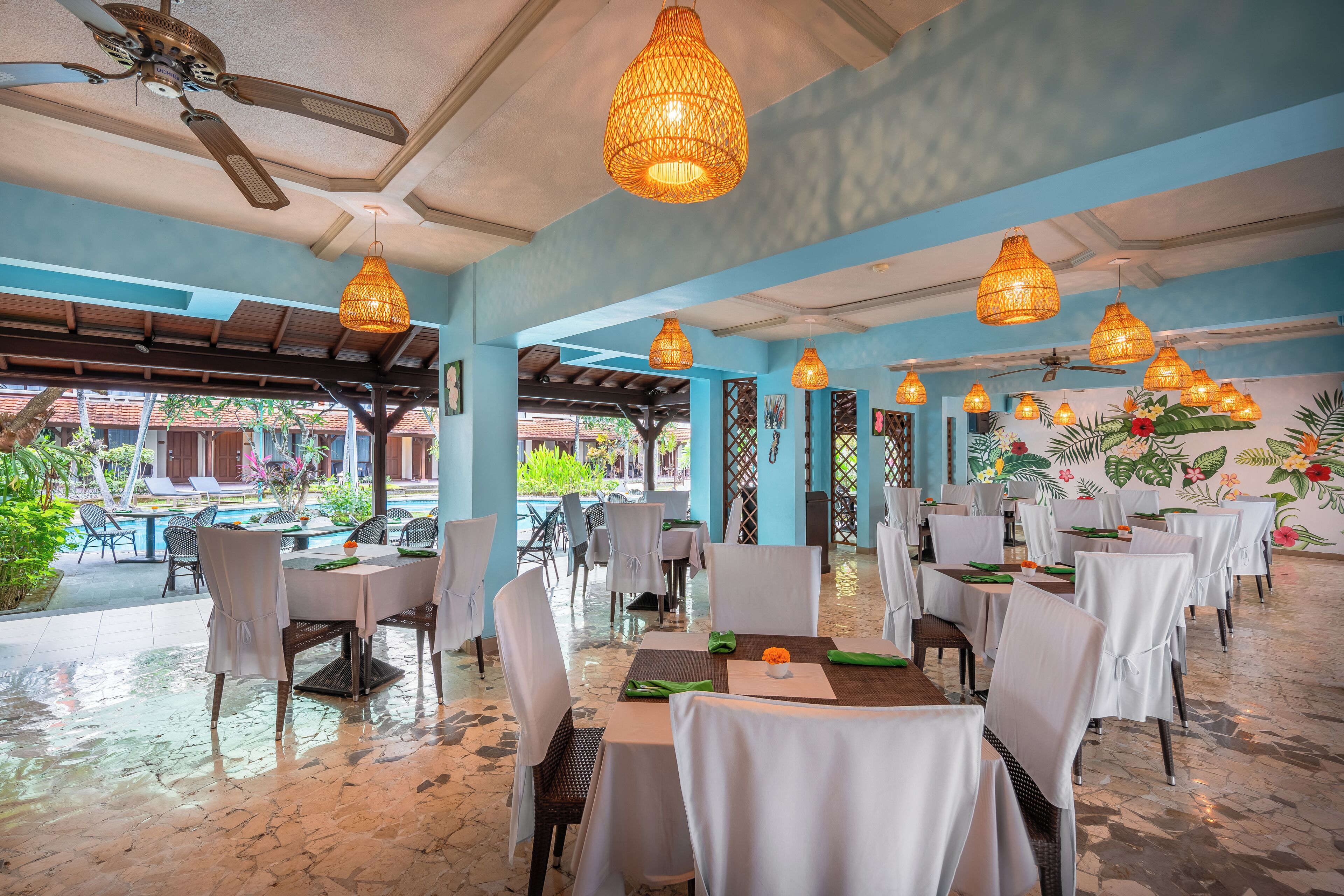 Un restaurant avec des tables dressées pour le service, des ventilateurs de plafond et des lampes en osier suspendues.