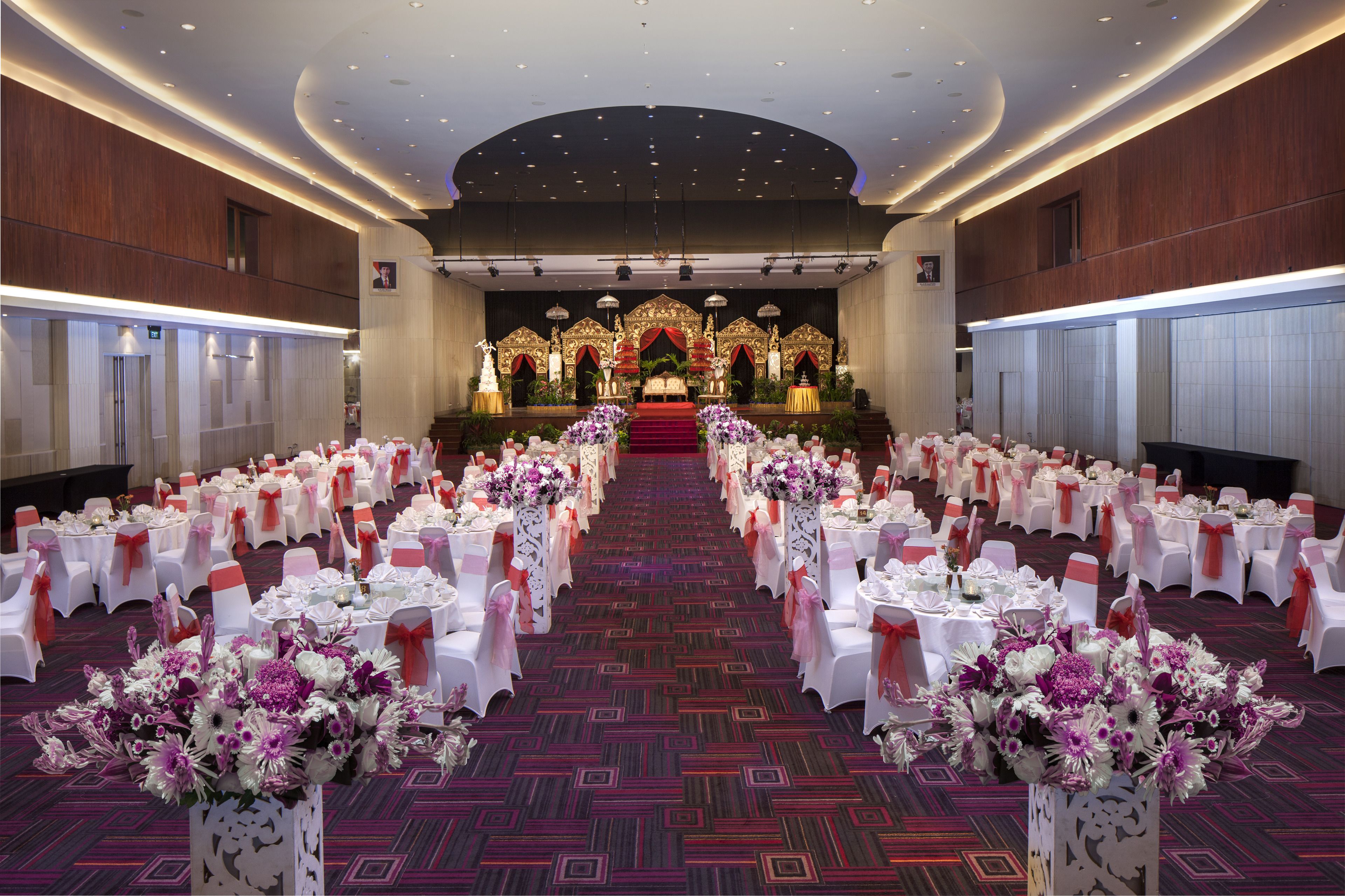 A banquet hall with rows of tables and chairs, a stage with floral decorations, and a patterned carpet.