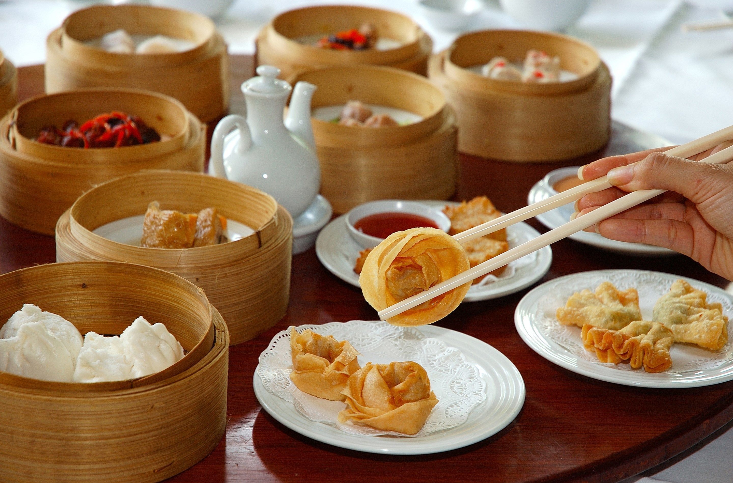 A variety of dim sum dishes in bamboo steamers and on plates, with a hand using chopsticks to pick up a dumpling.