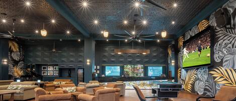 A spacious lounge area with multiple sofas, a pool table, and a bar area with a large screen displaying sports.