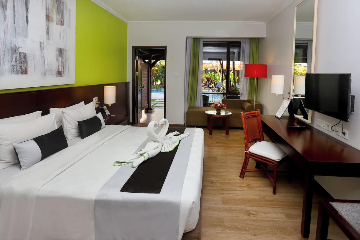 Ein Hotelzimmer mit einem aufgeräumten Bett, einer Couch, einem Schreibtisch mit Fernseher und Blick ins Freie.