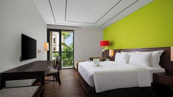 A hotel room with a bed, bedside table, lamp, TV, and a view of greenery through the window.