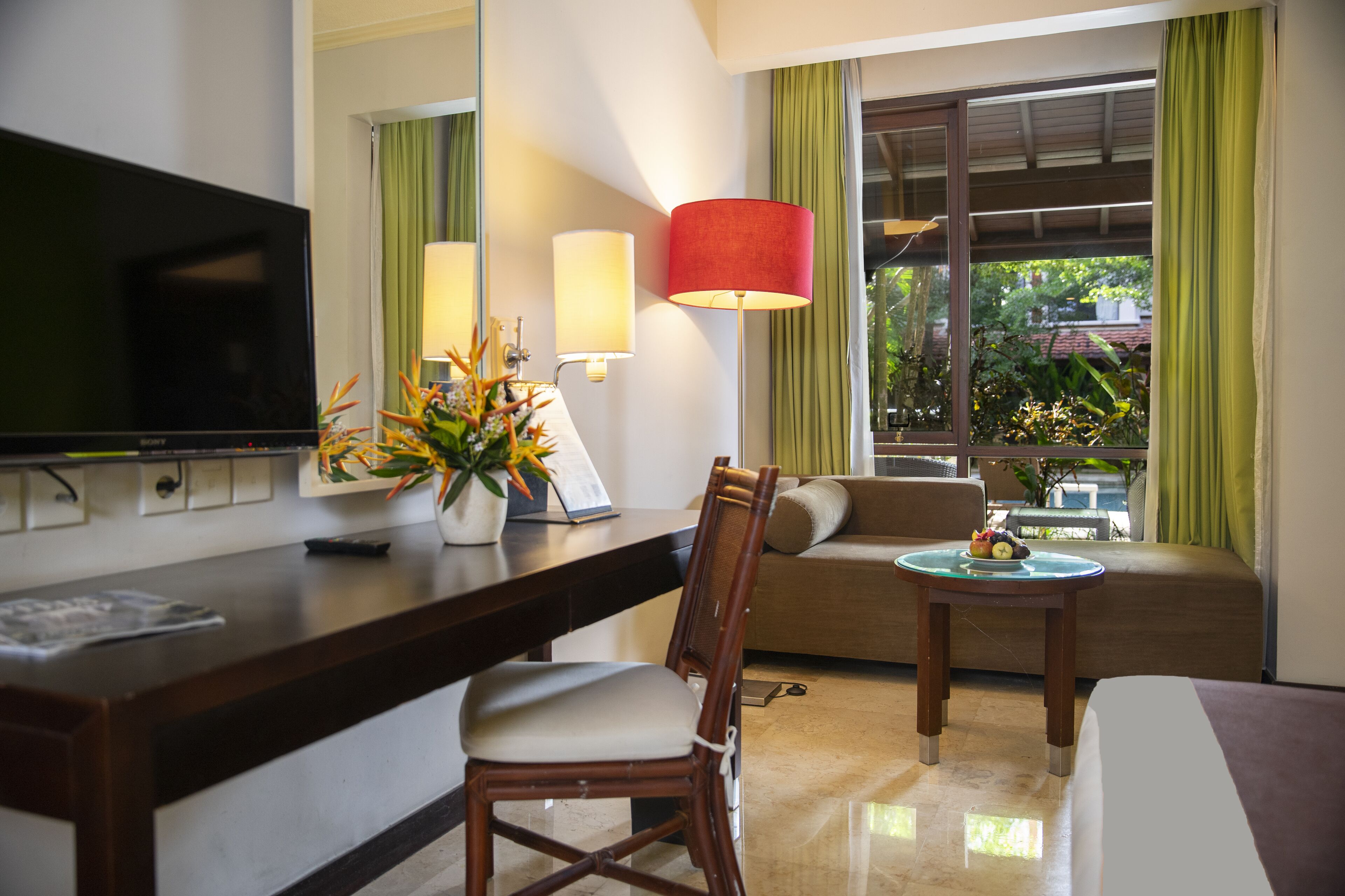 Una habitación de hotel con televisor, un escritorio con un florero, un sofá, una mesita de noche con lámpara y una ventana con cortinas.
