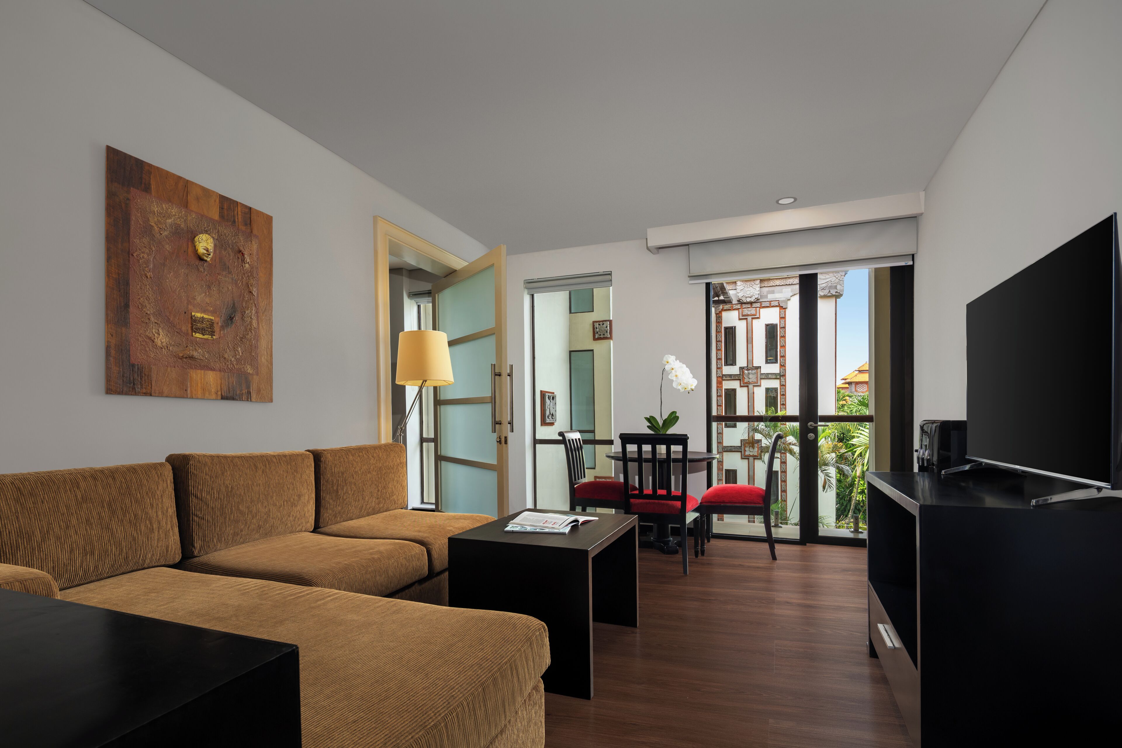 Una sala de estar moderna con un sofá, una mesa de centro y un televisor. Hay un balcón con vistas a edificios y plantas.