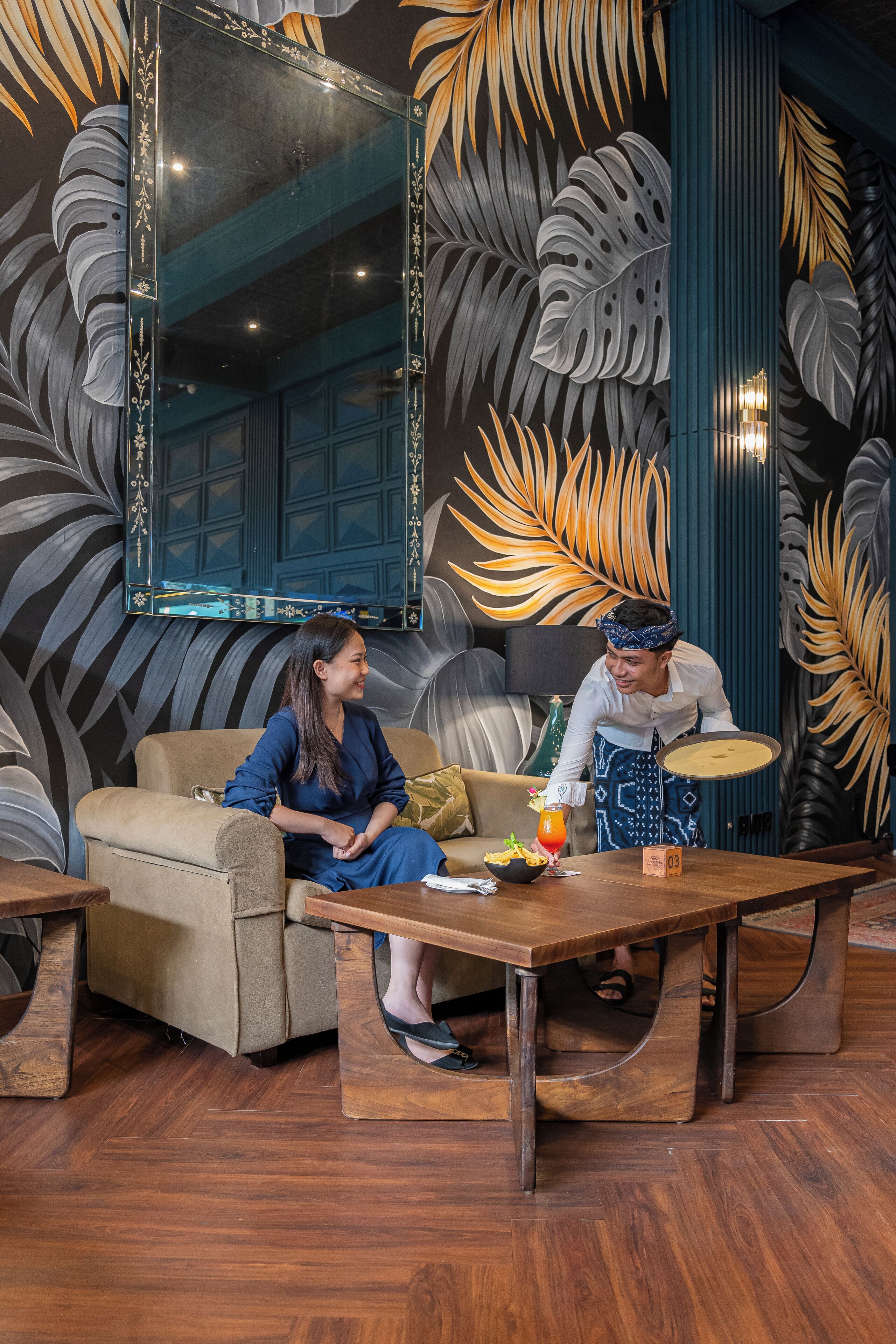 A room with a large mirror, a sofa, a wooden coffee table, and a decorative wall with leaf patterns.