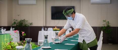Una persona preparando una mesa con cristalería transparente y platos en una habitación con mantel verde, sillas blancas y plantas en macetas.