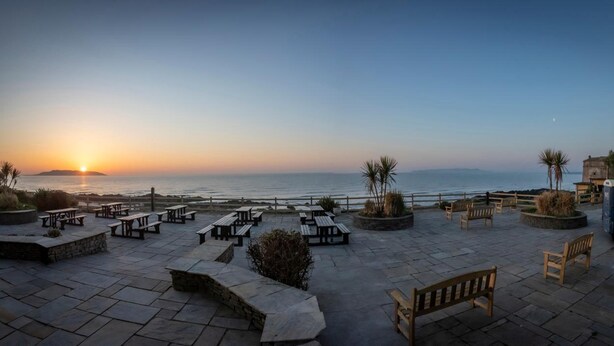 A seaside patio with stone tables and benches, a parked van, and a view of the sunset over the ocean. - Shoreline Hotel (Donabate)