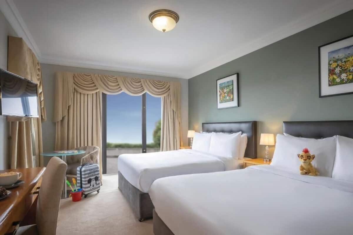 A hotel room with two beds, a desk, a chair, a suitcase, and a window with curtains.
