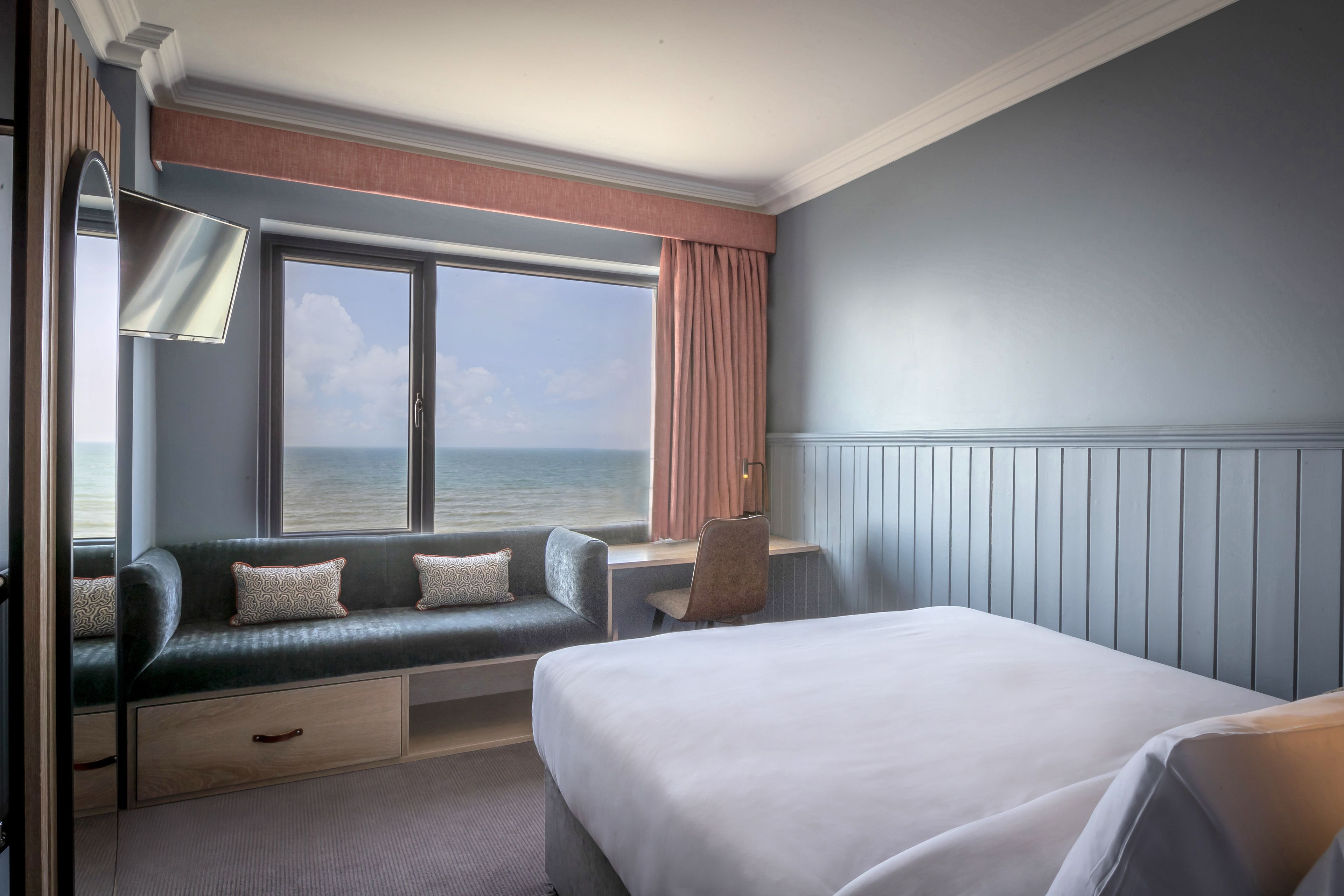 A hotel room with a bed, a seating area with a view of the sea, a desk, and a lamp.