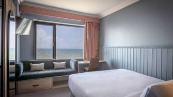 A hotel room with a bed, a seating area with a view of the sea, a desk, and a lamp.