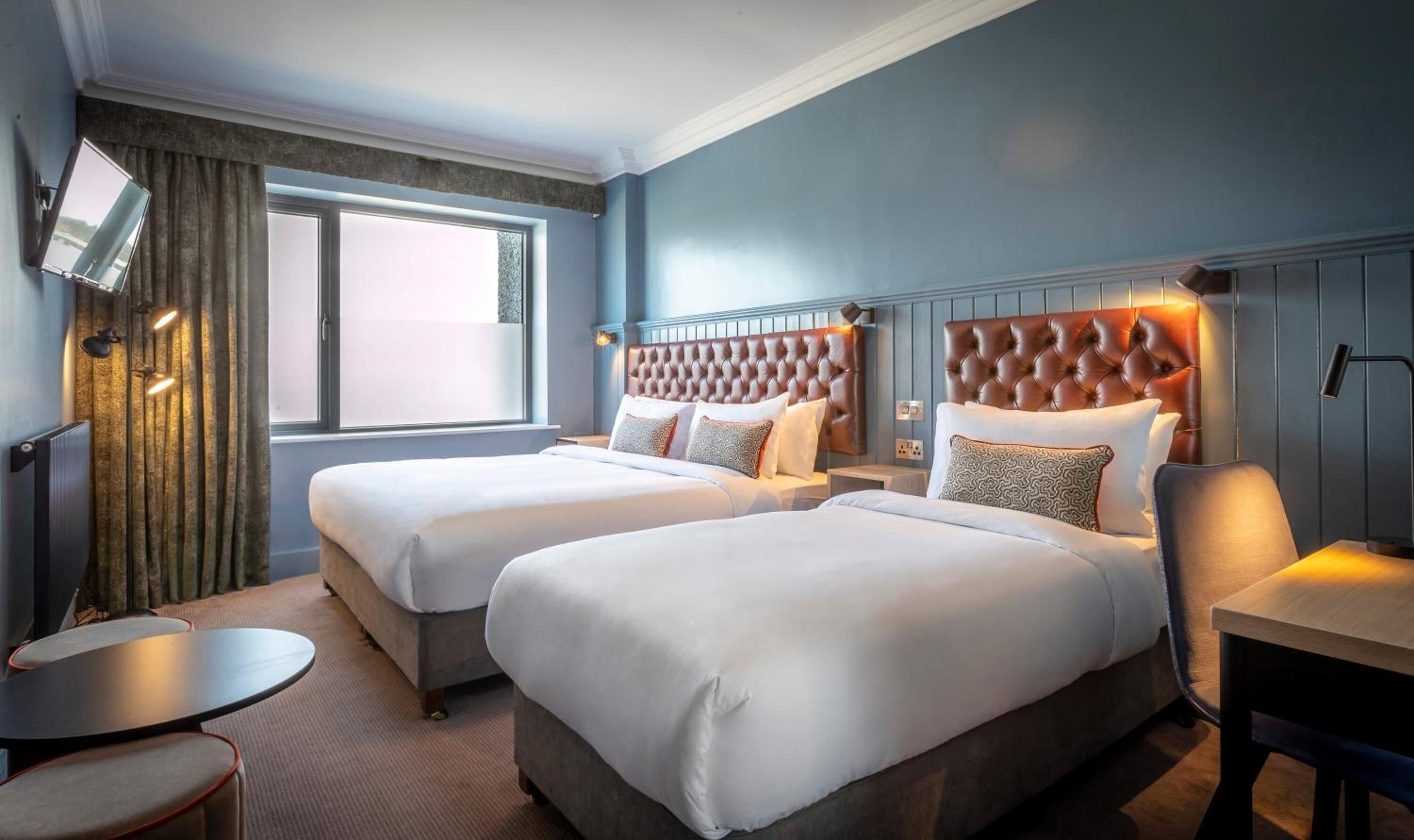 Habitación de hotel con dos camas, una mesa redonda pequeña, un escritorio y un ventanal con cortinas.