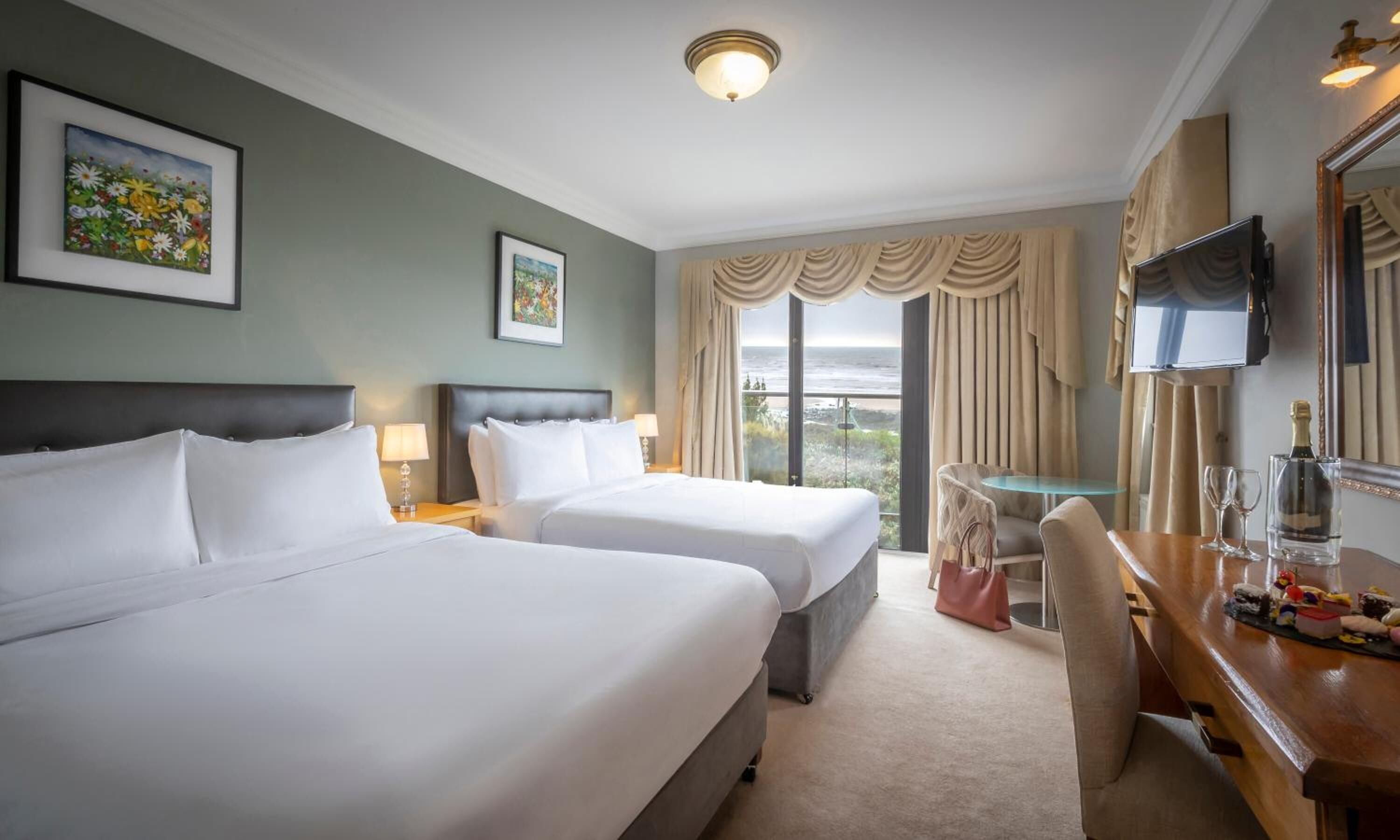 A hotel room with two beds, a TV, a desk with a bottle and glasses, and a view of the ocean.