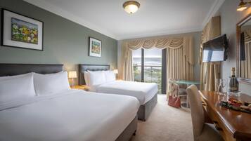 A hotel room with two beds, a TV, a desk with a bottle and glasses, and a view of the ocean.