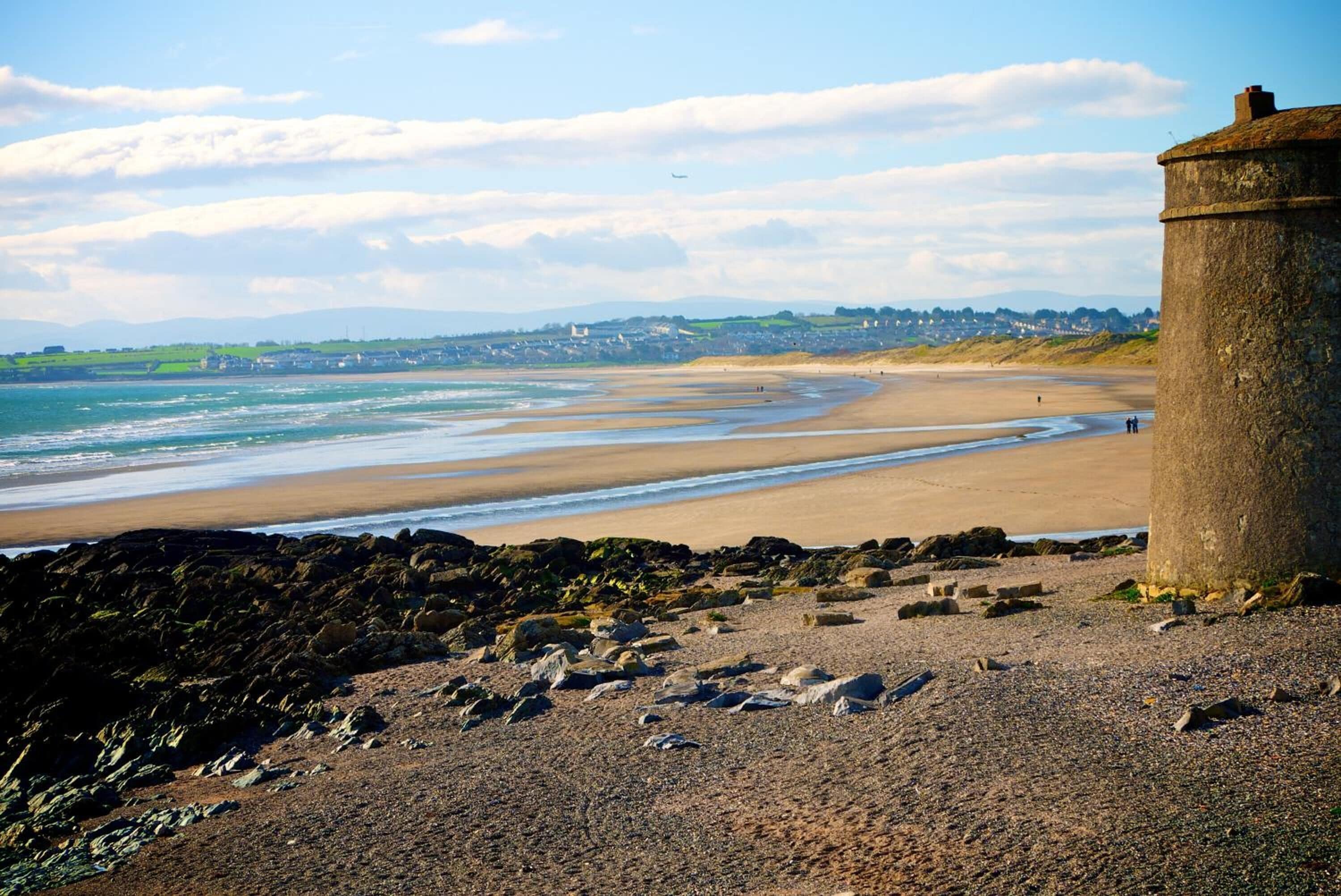 Un paysage côtier avec une plage de sable, un premier plan rocheux et une grande structure en pierre.