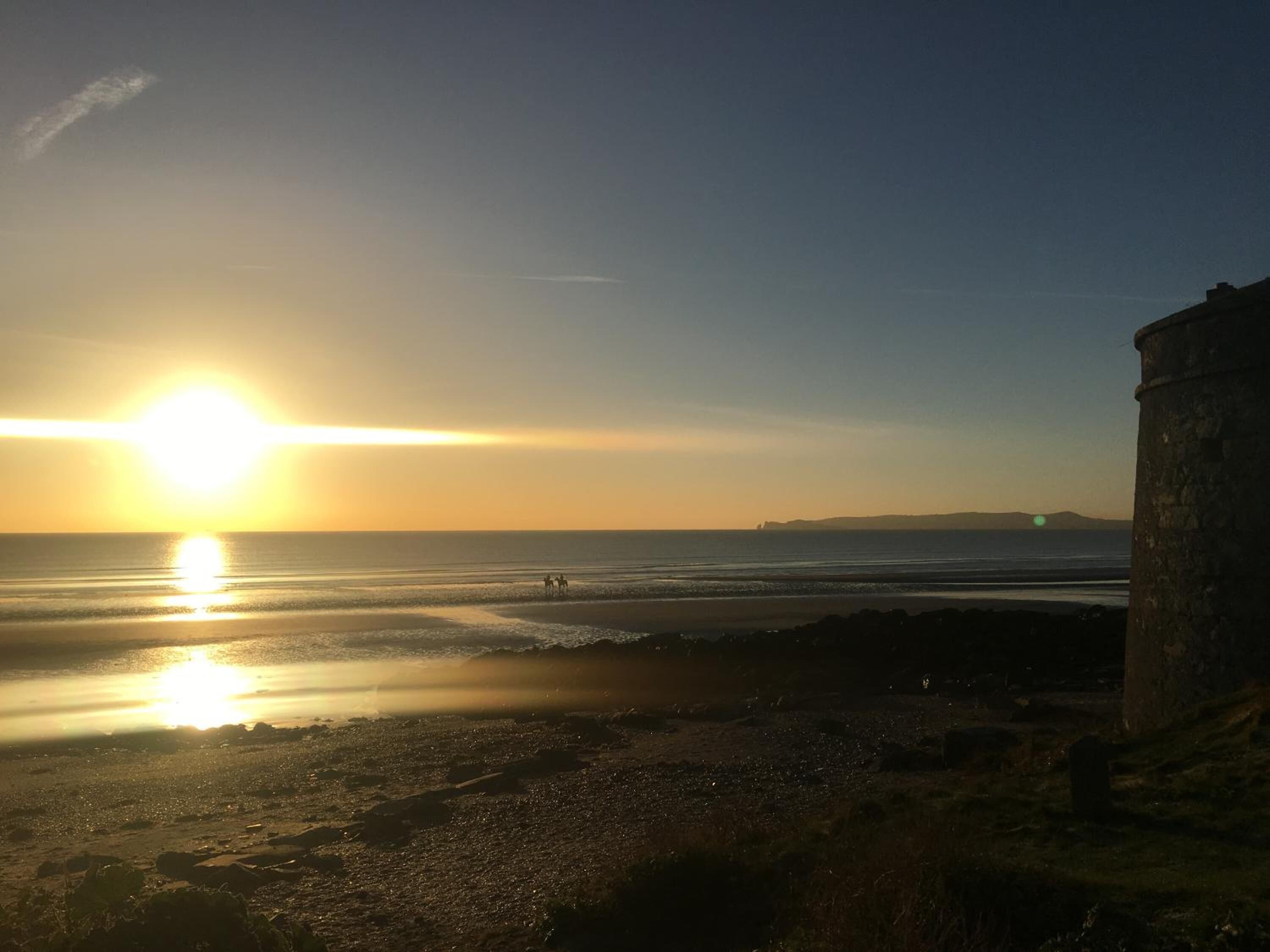 A beach scene with a sunset, a stone structure on the right, and a few people in the distance.